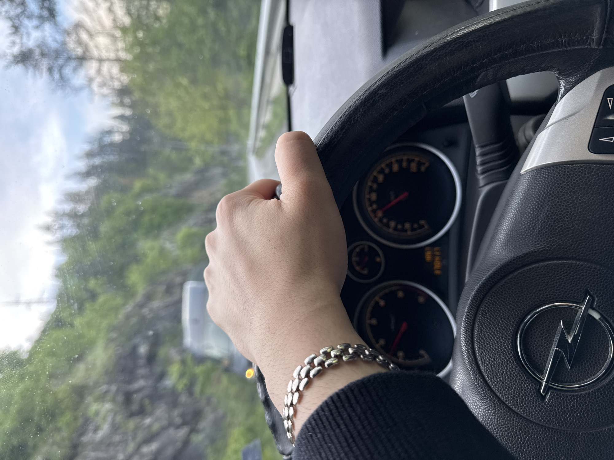 A photograph taken from inside a moving vehicle shows a hand gripping a black steering wheel with an Opel logo, suggesting the vehicle is an Opel. A silver bracelet adorns the driver's wrist. The vehicle's dashboard with gauges is visible in the background. Outside the passenger window, a blurry landscape of trees and a building is visible. The location is Taran, Bulgaria.