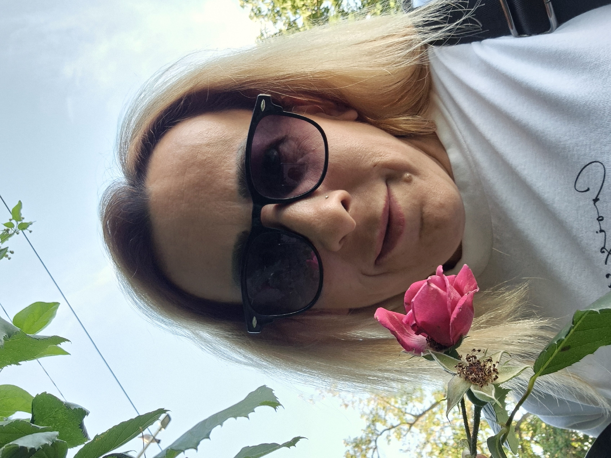 This is a low-angle, close-up, vertical, first-person perspective selfie of a person wearing black sunglasses and a white t-shirt. The person has blonde hair and is outdoors, with greenery and a bright sky visible in the background. A pink rose is held in the foreground, with some of its petals beginning to wilt and the center showing seed pods. The person's face is partially obscured by the sunglasses and the angle of the shot. There appears to be some light text or a graphic on the t-shirt, but it is not clearly legible. The overall impression is a casual, sunny day.