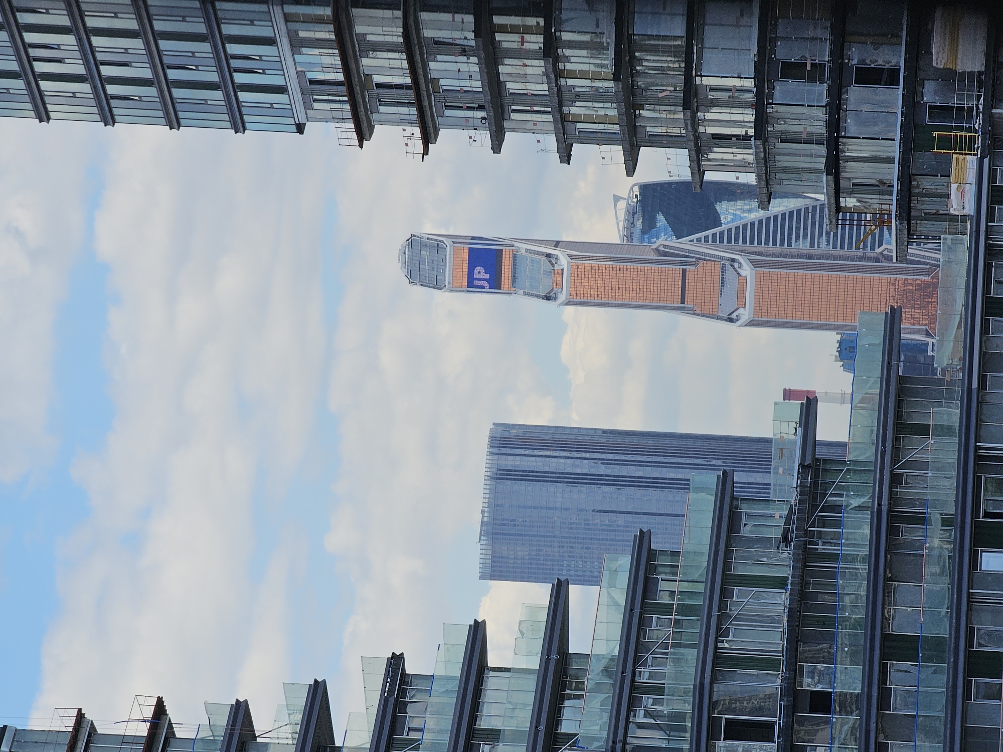 The image shows a view of the Moscow City business district on a cloudy day. In the foreground, on the left and right sides, are unfinished modern residential buildings under construction. These buildings feature multiple floors with exposed concrete frames, scaffolding, and glass balconies. The facades are partially covered with protective plastic sheeting.
In the middle ground, several prominent skyscrapers are visible. The most striking is the Neva Towers complex, specifically the skyscraper with a copper-colored facade, which prominently displays the blue "JP" logo on a digital screen. To its left and slightly behind is another skyscraper with a blue glass facade, part of the same complex. Further to the left, a taller, narrower skyscraper with a grey, ribbed facade stands out. The sky is filled with white and grey clouds against a pale blue background. The overall scene depicts a developing urban landscape with a mix of construction and completed modern architecture.