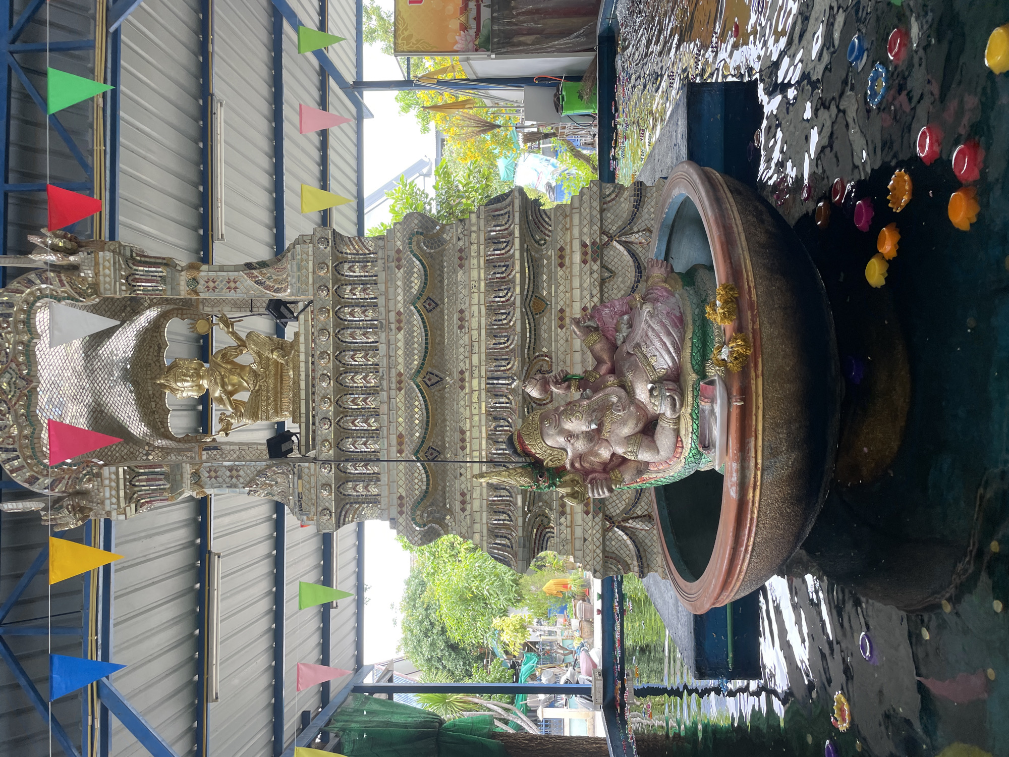 In Tambon Na Chom Thian, Thailand, a scene unfolds within an open-air structure featuring a corrugated metal roof supported by blue metal beams. Colorful triangular flags in blue, yellow, pink, and green are strung across the beams, adding a festive touch. The focal point is a large, ornate tiered structure, heavily decorated with mirrored mosaic tiles. Atop this structure sits a golden deity, possibly a Buddha or Hindu god, with multiple arms.
Positioned in front of this elaborate shrine, within a large, circular, reddish-brown basin filled with dark water, is a statue of the elephant-headed deity Ganesha. The Ganesha statue, rendered in a pinkish hue with green and gold accents, is seated and holds various objects in its hands. The basin itself rests on a submerged blue platform.
The surface of the water in the basin and the surrounding area is dotted with numerous small, colorful floating objects that resemble flowers or lotus blossoms, predominantly in shades of yellow, orange, red, pink, and purple. The water reflects the surrounding environment and the structures, creating a shimmering effect. Beyond the immediate shrine area, glimpses of lush green trees, buildings, and a clear sky suggest an outdoor setting. The overall impression is one of a spiritual or cultural site, likely a temple or shrine, decorated for an occasion.