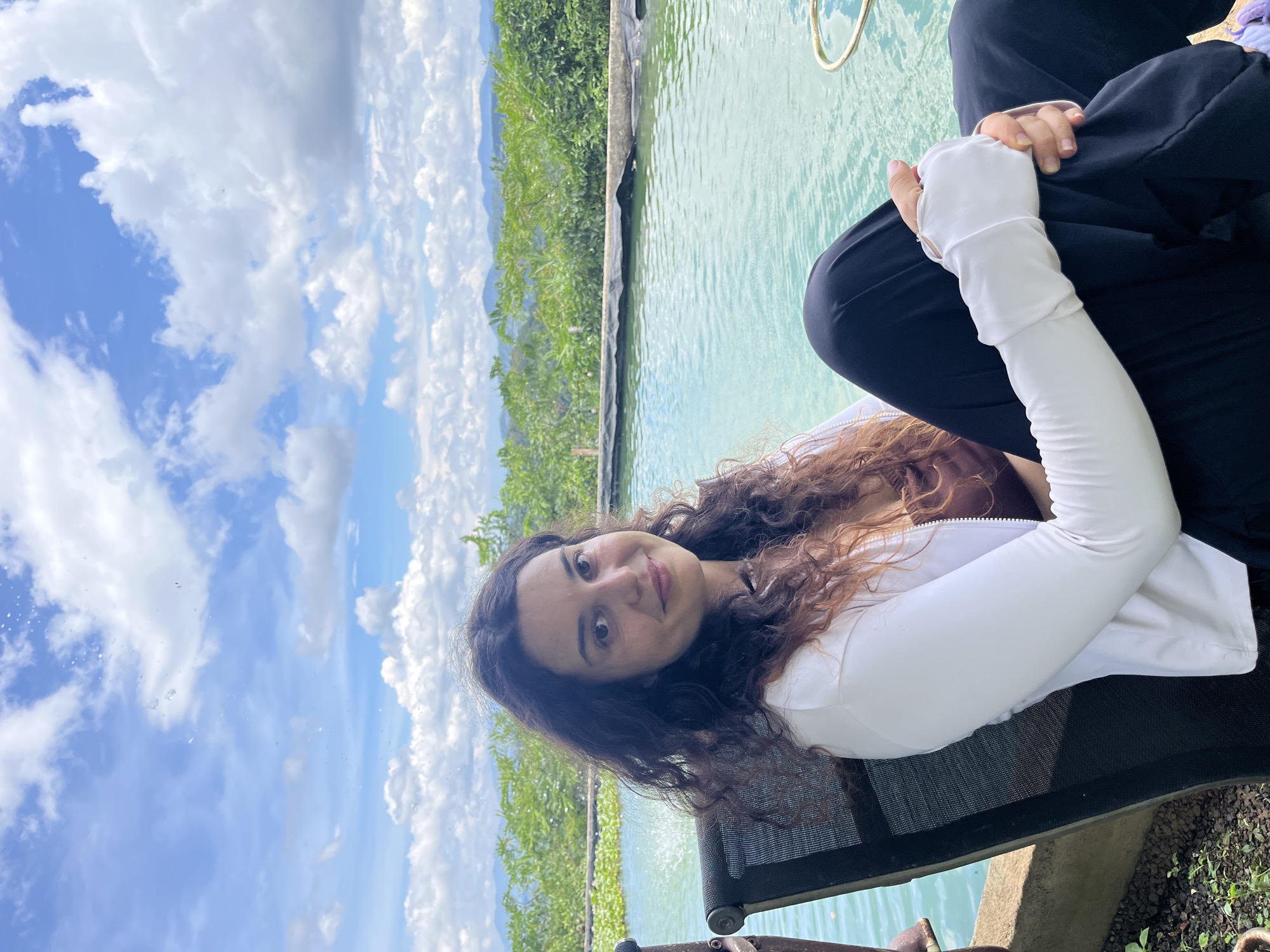 A woman with long, curly brown hair is sitting in a black chair next to a pool of turquoise water. She is wearing a brown top and a white jacket. Her hands are resting on her lap, and she is looking at the camera with a slight smile. The background shows a lush green landscape with mountains in the distance and a blue sky with white clouds. The scene appears to be outdoors in a tropical or subtropical region. The location is identified as Sơn Điền, Vietnam.
