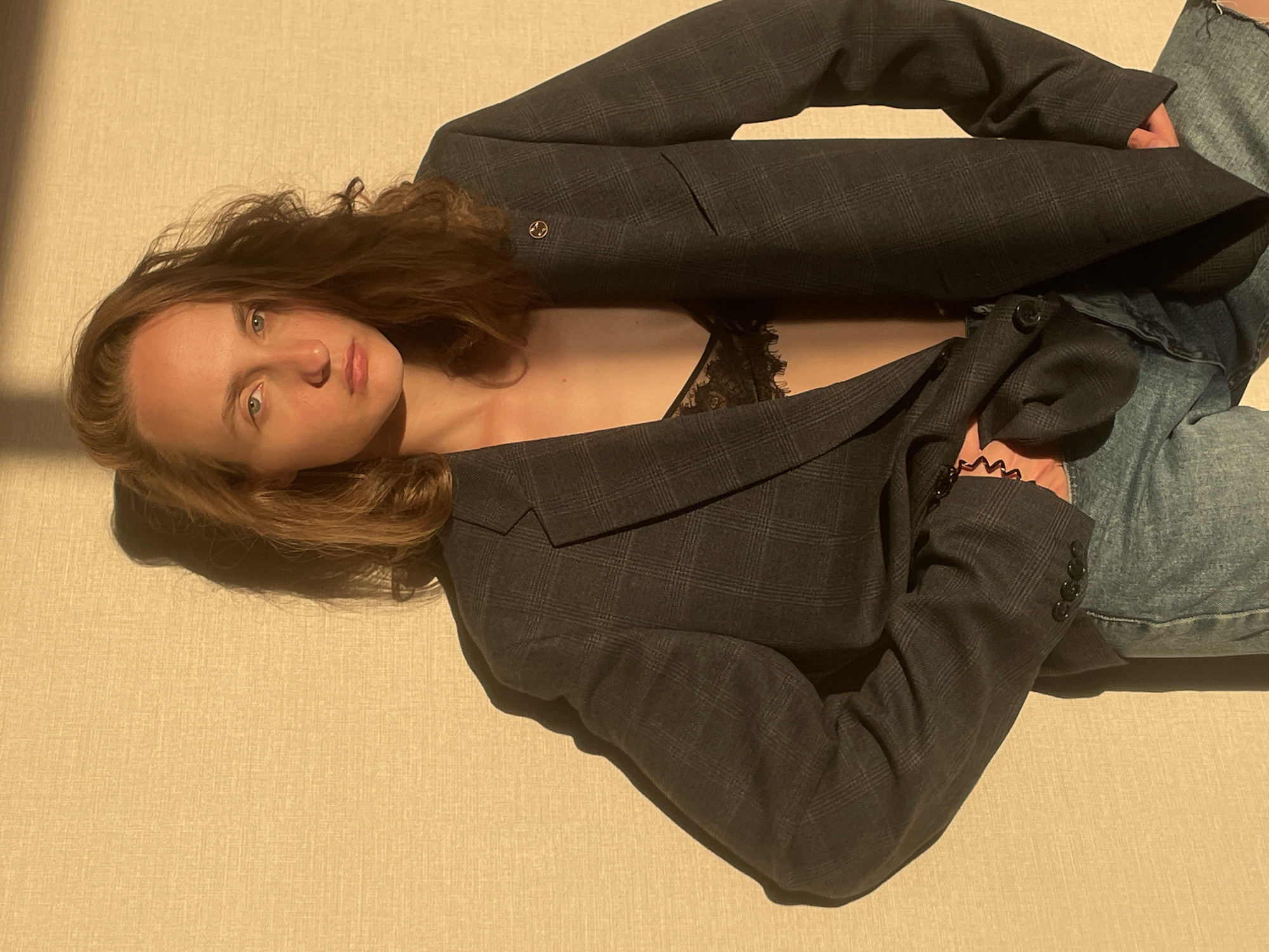 A woman lies on a beige textured surface, possibly a bed or couch. She is oriented diagonally, with her head resting on the left side of the frame. She has long, reddish-brown hair and is wearing a black lace bralette under a dark grey plaid blazer. Her jeans are visible on the right side of the frame. Her expression is neutral, and she appears to be looking directly at the viewer. The lighting is warm and creates strong shadows on the background. The exact location in the city of Подшивалово, Russia, is not evident from the image.