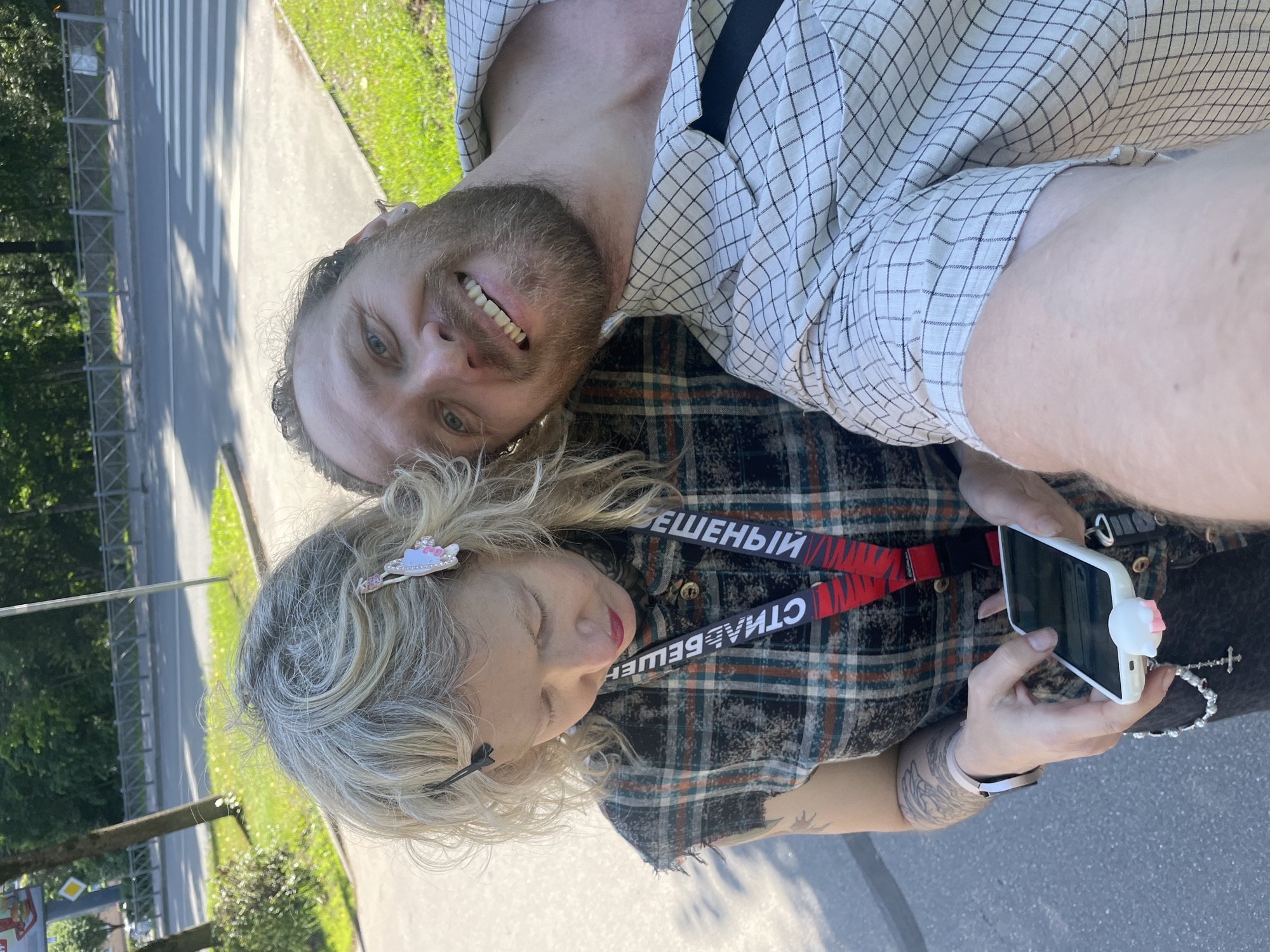 This is a candid, slightly blurry selfie taken outdoors. The camera is held by a woman with blonde, curly hair, who is looking down. She is wearing a plaid shirt and a lanyard with red and black straps that has text on it. A white phone with a pink cat-shaped attachment is visible in her hands. To her right is a man with a beard and blue eyes, smiling at the camera. He is wearing a checkered shirt. In the background, a street with a pedestrian crossing and greenery can be seen, suggesting a daytime setting on a sunny day. The overall mood appears to be relaxed and casual. The visible text on the lanyard appears to be in Cyrillic, and includes the words "БЕШЕНЫЙ" and "СТИЛЬ".