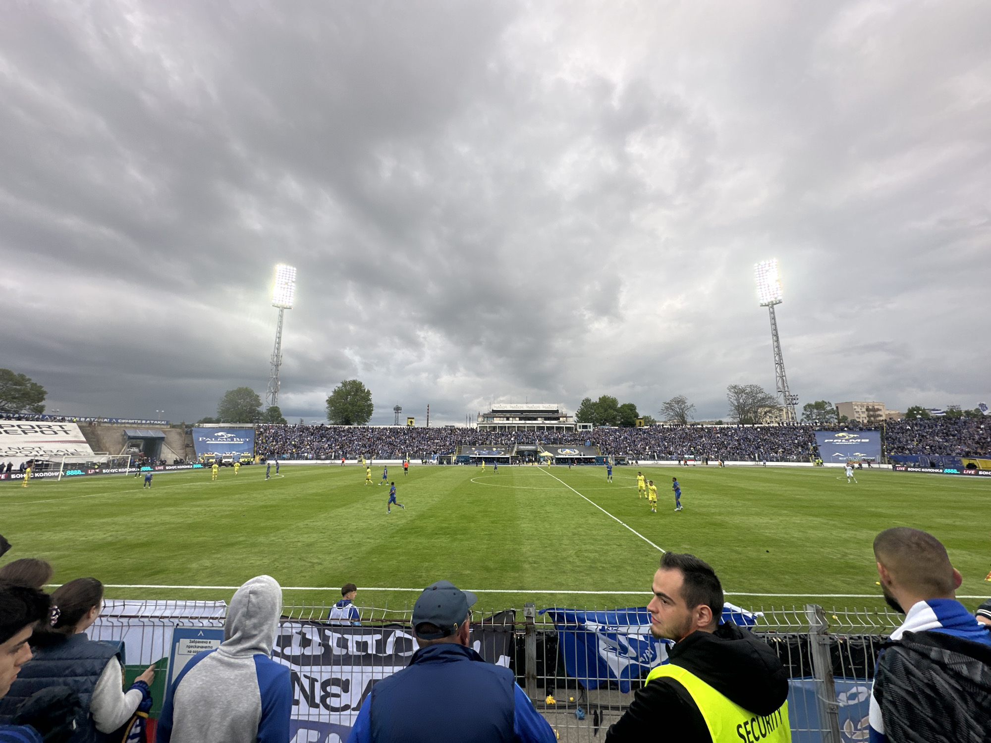 The image shows a soccer game in progress at a stadium. The stadium is filled with spectators. The players are wearing blue and yellow uniforms. The field is green. In the foreground are several spectators standing near a metal railing. One of the spectators is wearing a yellow security jacket. The sky is overcast. The location is likely in Sofia, Bulgaria, based on the Cyrillic text in the stadium advertisements. There are floodlights illuminating the field.
