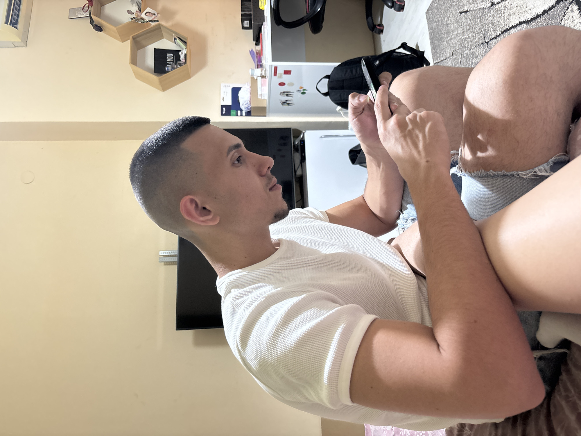 This image captures an indoor scene in a residential dwelling in Plovdiv, Bulgaria. A male, appearing to be in his late 20s to early 30s, is seated and viewed in profile from the right. He has a very short, faded haircut and a slight goatee. He is wearing a white short-sleeved waffle-knit t-shirt and light blue distressed denim shorts or jeans. His arms are bent, and both hands are holding a black smartphone, with his right index finger actively touching the screen. He is focused intently on the device. The bare leg of another individual is partially visible in the foreground, positioned in front of the male's legs, suggesting two people are seated closely together on a surface covered by a patterned brown or maroon blanket or sheet.
The room features light beige walls. In the background, to the left, a large, dark flat-screen television is visible on a stand. To the right of the television, a white mini-refrigerator is situated, displaying several magnets or stickers, including stick figures. Above the mini-refrigerator, a white desk holds various objects, including a box labeled "Trust." A black office chair with red accents is partially visible next to the desk. Two hexagonal wooden shelves are mounted on the wall above the desk; the lower shelf contains a black object with the text "4 YOU." A white air conditioning unit is installed high on the wall in the upper right portion of the frame. A black backpack rests on the floor between the desk and the office chair. The floor is covered by a light-colored, patterned rug. Artificial lighting illuminates the space.