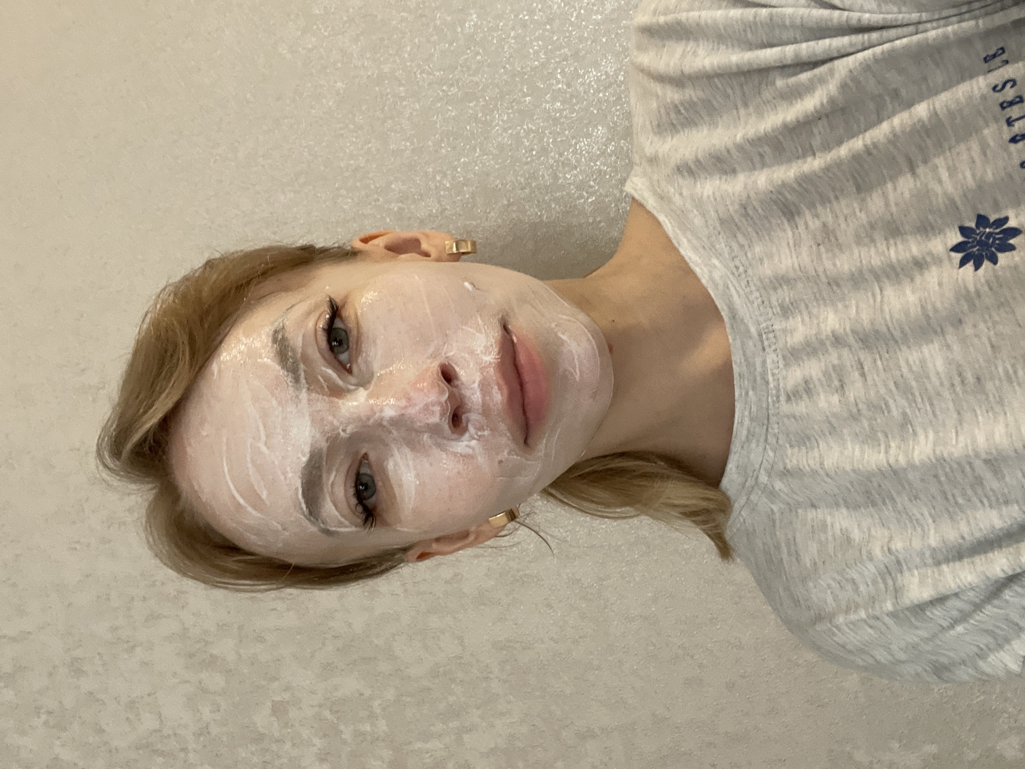 A fair-skinned woman with blonde hair is shown from a close-up, slightly angled, portrait perspective. She is wearing a white facial mask. She is wearing a gray t-shirt with a blue flower emblem and part of text "ATES LE" visible. She has gold earrings. The background is a textured off-white wall. This is in the city of Perm, Russia.