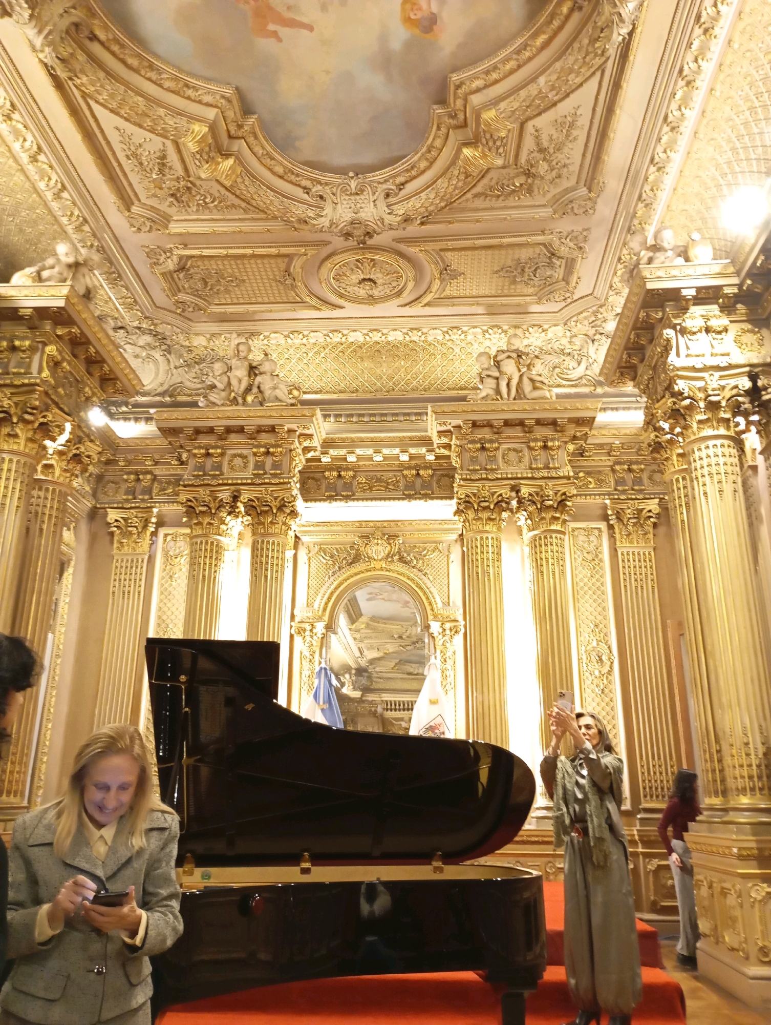 The image depicts the interior of the Salon Dorado at the Teatro Colón in Buenos Aires, Argentina. The highly ornate room features gilded stucco work, elaborate moldings, and painted ceiling frescoes depicting cherubic figures. A grand piano is positioned centrally on a red carpeted stage.
In the foreground, a woman with blonde hair, wearing a grey tweed jacket and a light-colored shirt, is looking down at her phone, which she holds in both hands. To her right, a woman with long blonde hair, dressed in a long, flowing green garment and a scarf, is standing and holding up a smartphone to take a photograph. Further to the right, a person in a dark top and light-colored pants is visible in the background. The opulent surroundings suggest a formal or artistic venue.
