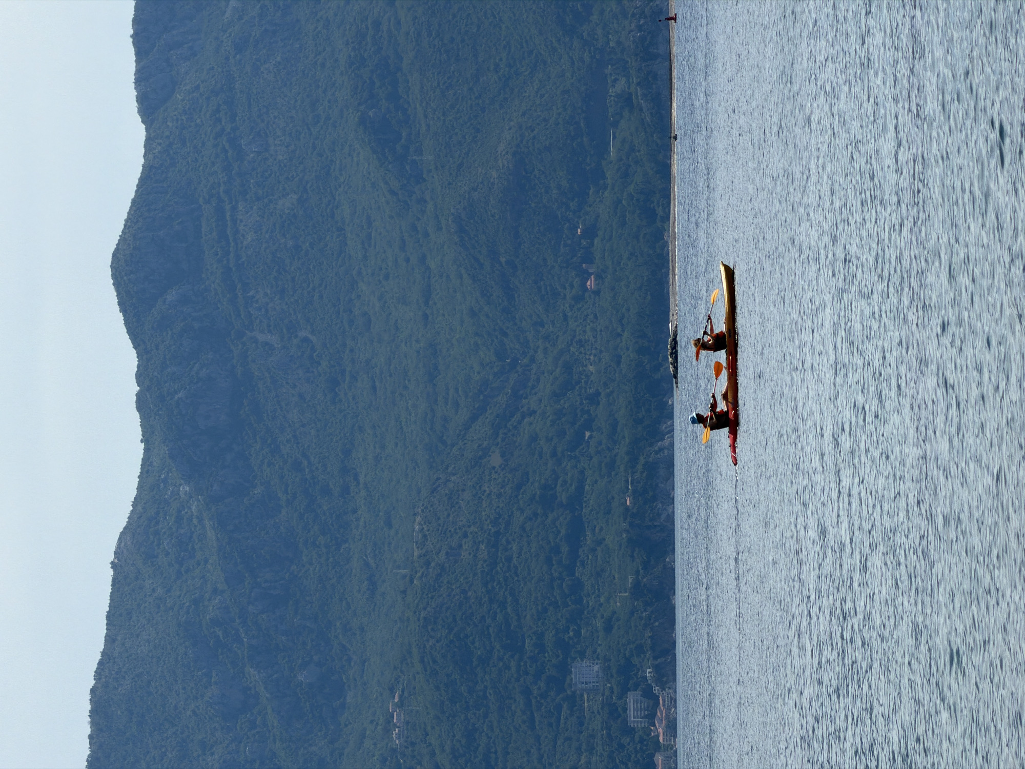 Two individuals are kayaking on the Adriatic Sea near Budva, Montenegro. The scene includes a long, narrow kayak occupied by the two people, mountains covered with dense green vegetation, and the water reflecting the sky. Buildings are visible in the distance near the shoreline at the base of the mountain.