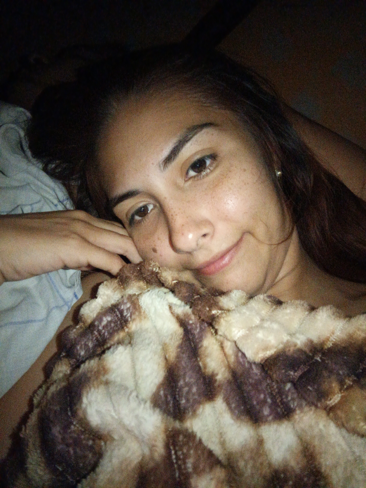 A woman with freckles is lying in a bed covered by a patterned blanket. Her head is resting on a pillow. The environment appears dimly lit. There is no indication of the location being Juan Griego, Venezuela.