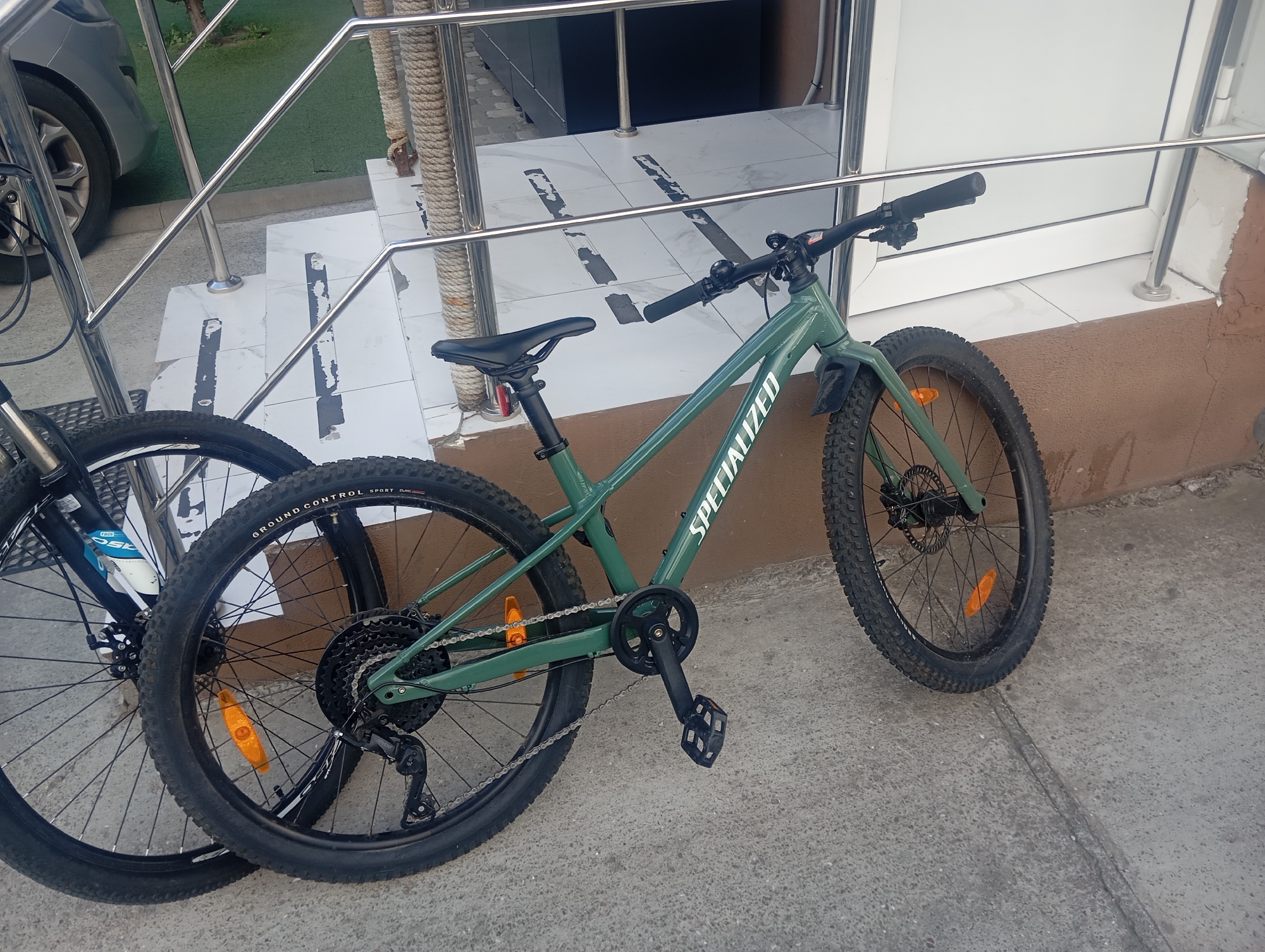 In Sofiivska Borshchahivka, Ukraine, two bicycles are parked outdoors on a concrete surface next to a building. The foreground features a green Specialized mountain bike with knobby tires, disk brakes, and a chain. To the left of this bicycle, another bicycle's rear wheel and part of the frame are visible, with a black tire and spokes. The bicycles are positioned near a set of white tiled stairs with grey accents, leading up to a building entrance. A silver car is partially visible in the background on the left. The environment appears to be a residential or commercial area, with a metal railing and a rope-wrapped post alongside the stairs. The overall scene is static, with no human activity depicted.