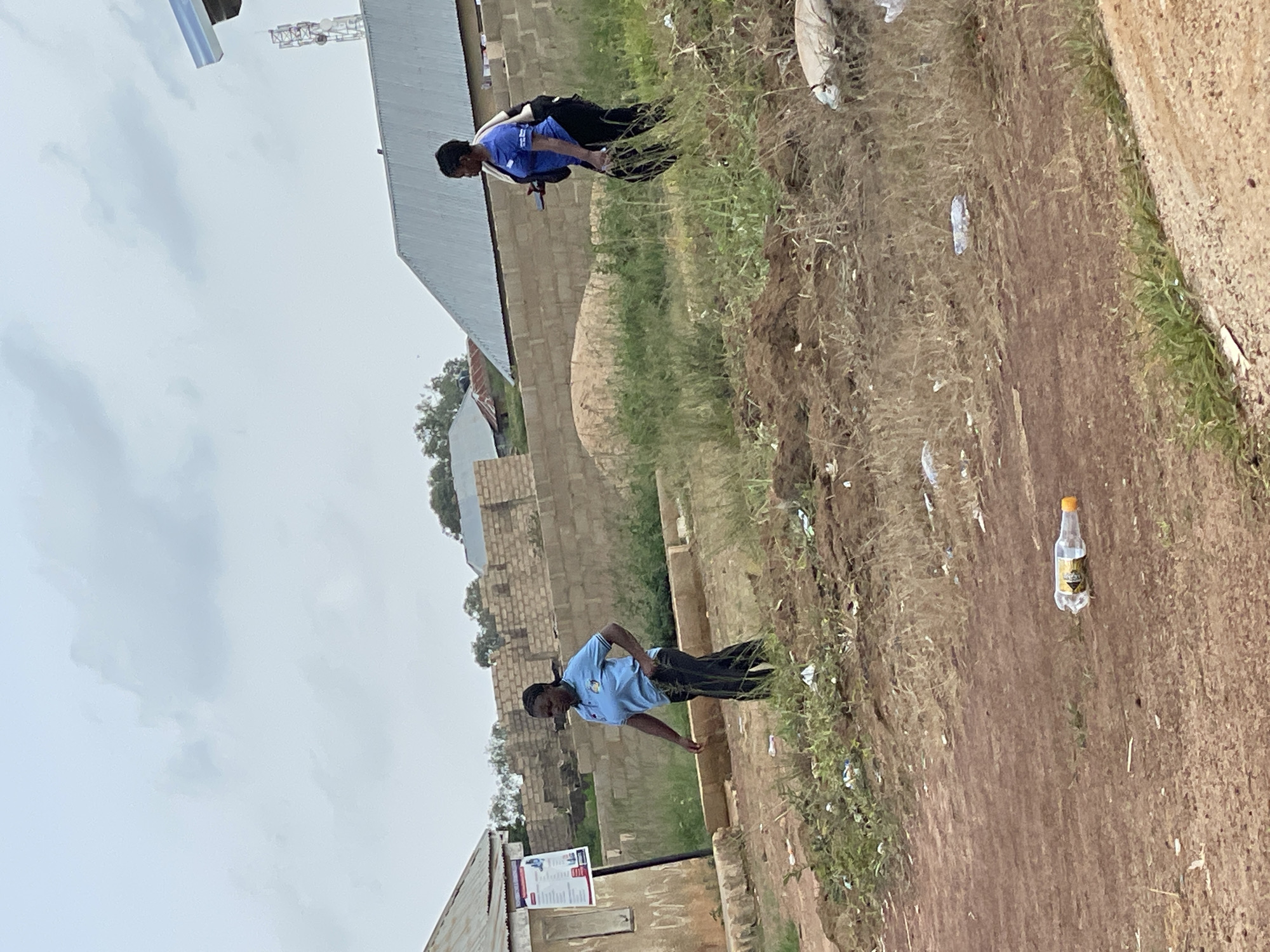 The image shows two individuals in a semi-urban or rural setting in Durumi, Nigeria. One person is standing and facing towards the right side of the frame, wearing a blue polo shirt and dark pants, with a backpack. This individual appears to be looking down or to the side. Further up and to the left, another person is visible, also wearing a blue shirt and dark pants, with a backpack, in a similar pose, looking away from the camera.
The scene is characterized by a dirt path or road with sparse vegetation and some scattered debris, including plastic bottles. In the background, there are buildings constructed from light-colored bricks, and a structure resembling a metal roof. A cell tower or similar industrial structure is visible in the distance. The sky is overcast, suggesting a cloudy or possibly rainy day. The overall impression is of an outdoor environment, possibly a community area or the outskirts of a settlement.
There are no specific events or activities clearly depicted beyond the presence of the individuals. The time of day is not distinctly indicated, but the lighting suggests it is daytime. Emotions are not discernible from the image. A piece of paper or sign with text is partially visible in the lower left portion of the frame, but the text is not legible.
