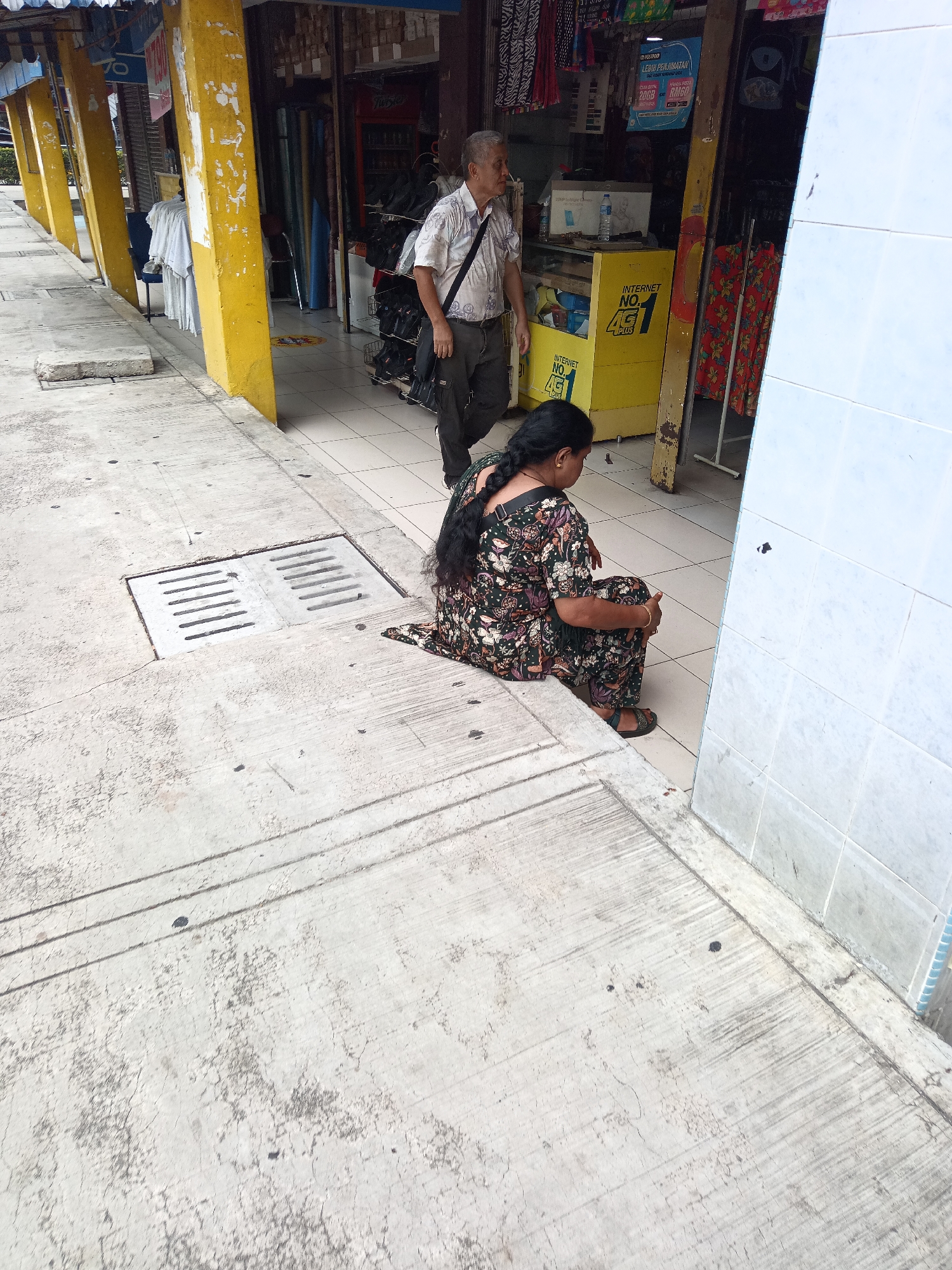 An overhead outdoor shot shows a woman with dark hair, wearing a floral print dress and sandals, sitting on a concrete sidewalk with her knees drawn up. She is facing a white tiled wall and a drain grate. Behind her, a man in a white short-sleeved shirt and dark pants, carrying a black bag over his shoulder, walks past a shop with yellow pillars and displays of merchandise. The location appears to be a commercial street in Kuala Lumpur, Malaysia, possibly in a market area.