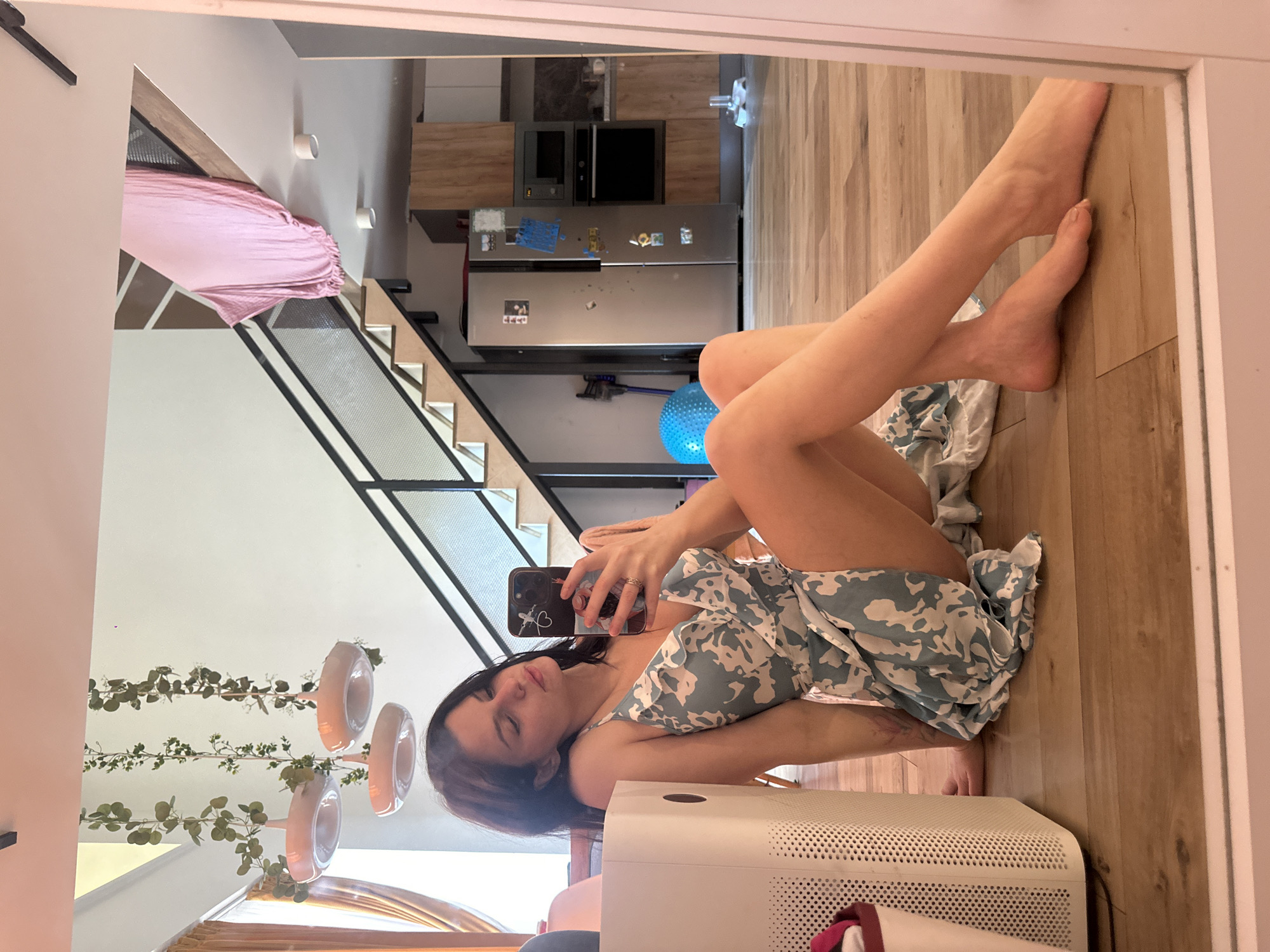 A young woman with dark hair is sitting on a wooden floor, taking a mirror selfie with her smartphone. She is wearing a light blue and white patterned slip dress. To her left, there is a white mesh laundry hamper. In the background, a staircase with black railings and wooden steps leads up to a landing. A pink fabric is draped over the railing on the upper level. Further back, a kitchen area is visible with wooden cabinets, a stainless steel refrigerator, and a microwave. The room is well-lit, with several pendant lights hanging from the ceiling and greenery draped around them. The scene appears to be indoors, possibly in a modern apartment.