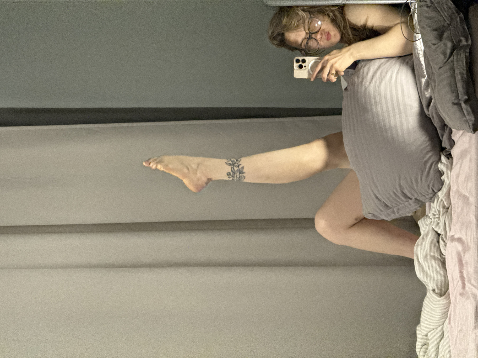 A woman with glasses and a phone takes a selfie in a mirror, partially obscured by bedding. A leg extends into the frame, featuring a floral tattoo above the ankle. The location and details of the city of Chaikovsky, Russia, are unknown.