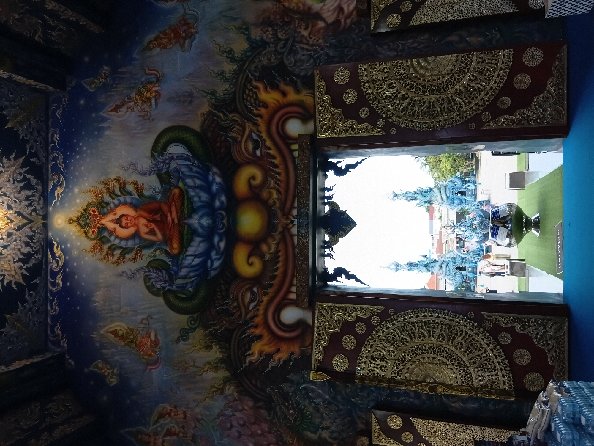 This image depicts the entrance to the Wat Rong Suea Ten, also known as the Blue Temple, in Chiang Rai City Municipality, Thailand. The focal point is a highly ornate mural above the doorway, featuring a seated Buddha figure surrounded by mythical serpents and celestial beings against a backdrop of a starry sky and misty mountains. The doorway itself is framed by intricate, dark red panels adorned with elaborate gold-filigree carvings. Through the open doorway, a view of the temple's exterior is visible, showcasing striking blue sculptural elements, including dragon-like formations and stupas, set against a bright sky. Several individuals are seen standing outside the temple. In the foreground, on the left side of the image, stacks of bottled water and bags of rice are placed, suggesting they might be offerings or for sale. A silver-toned bowl on a pedestal sits on a green carpeted area in front of the doorway. The overall scene is a blend of interior architectural detail and a glimpse of the vibrant exterior of a significant religious site.