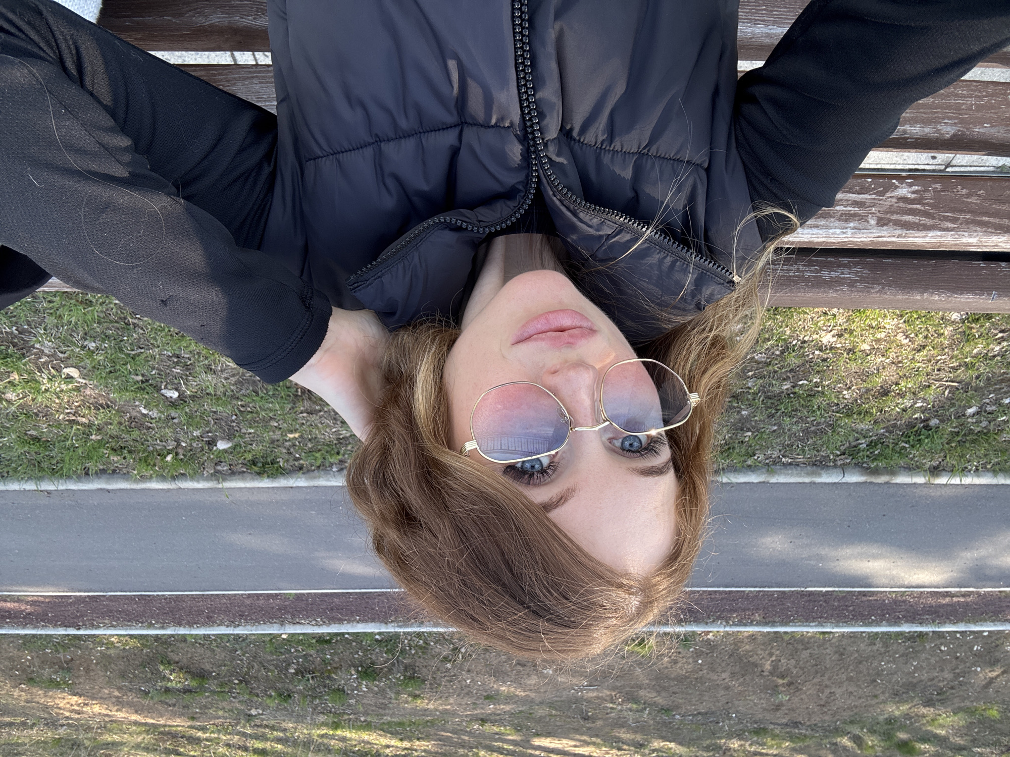 A woman with light brown hair and wearing gold-framed sunglasses is lying on a park bench. She has on a black puffer vest and a black long-sleeved shirt. The background shows a paved pathway, grass, and trees. The image appears to be taken in a park in Chaikovsky, Russia.