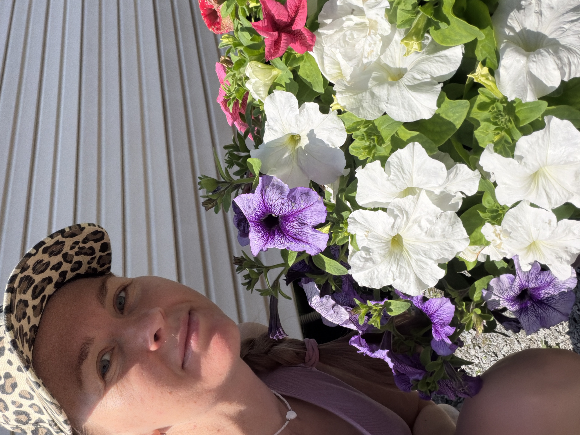 In Khekhtsir, Russia, a Caucasian woman with blonde hair, wearing a leopard-print baseball cap and a lavender top, is shown from the chest up, looking slightly to her left. She is partially obscured by a planter filled with vibrant petunias in shades of purple, white, and pink. The background consists of a light-colored, horizontally paneled wall. The woman has a pearl necklace with a heart-shaped pendant. Her blonde hair is styled in a braid secured with a pink band. The petunias are in bloom, with some flowers in focus and others slightly blurred, creating a shallow depth of field. The scene is illuminated by natural sunlight, casting shadows on the woman's face and the surrounding foliage.