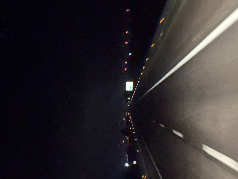 The image depicts a multi-lane highway at night, viewed from the perspective of a moving vehicle within the vicinity of Foggia, Italy. The dark asphalt roadway is delineated by a prominent solid white line on the immediate right shoulder and a dashed white line separating the current lane from an adjacent lane to the left. Numerous orange/amber rectangular reflective markers are interspersed along both the right and left edges of the roadway, receding into the distance. Ahead, white and blue headlights from oncoming or parallel traffic are visible on the far left, while red taillights of preceding vehicles appear as streaks due to motion blur. A rectangular, illuminated green road sign, with illegible white text, is situated in the middle distance above the right side of the highway. The sky above is uniformly dark and devoid of features. No individuals are visible. The overall image exhibits characteristics of low-light photography, including significant blur and grain.