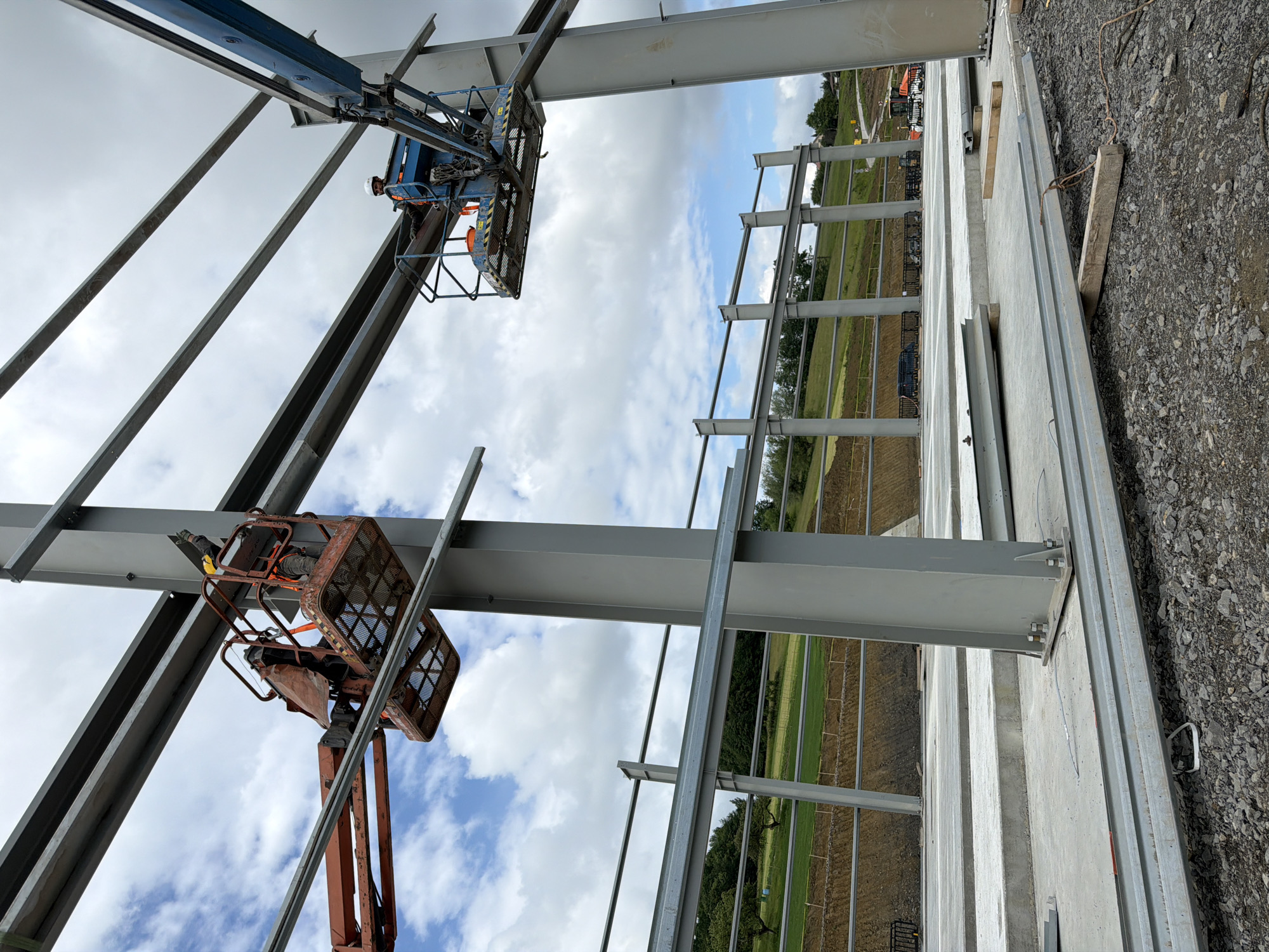 This image depicts a construction site in the city of Dobra, Poland, focused on the erection of a large steel-framed building. The foreground shows a concrete foundation with heavy steel beams laid out, some propped up on wooden planks. The midground features towering grey steel columns and beams, forming the skeletal structure of the building.
In the background, the landscape consists of green, rolling hills and trees, indicating an area on the outskirts of the city. Further back, construction vehicles, including what appears to be an orange excavator, are visible.
Two individuals are present in the scene, each operating from an elevated platform lift. One person, wearing a yellow hard hat and an orange vest, is in a red platform lift on the left side of the image, seemingly working on attaching a steel beam to a column. The other individual, in a white hard hat and an orange vest, is in a blue platform lift on the right, also engaged in construction work on the steel framework. The sky is partly cloudy with white cumulus clouds against a blue sky. The overall scene conveys an active construction project in progress.