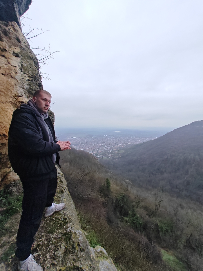 A male individual stands on a moss-covered, light brown rock ledge, overlooking the city of Shumen, Bulgaria, from an elevated position on the Shumen Plateau. The subject, appearing to be an adult male with short, light hair, is dressed in a black puffer jacket, a grey hooded sweatshirt, a grey scarf, black pants, and light-colored athletic shoes. His body is angled slightly left, while his head is turned to the right, observing the distant panorama. His hands are clasped together in front of his torso. To the immediate left is a steep, textured cliff face of a similar light brown rock. The valley below reveals the city of Shumen, characterized by numerous buildings with varied roofs, stretching across the landscape. Beyond the city, a body of water is faintly visible on the horizon. The slopes leading down from the plateau and the opposing hills are covered in a dense, bare deciduous forest, indicative of a colder season. The sky is uniformly overcast and light grey, contributing to a hazy atmospheric condition over the distant landscape.