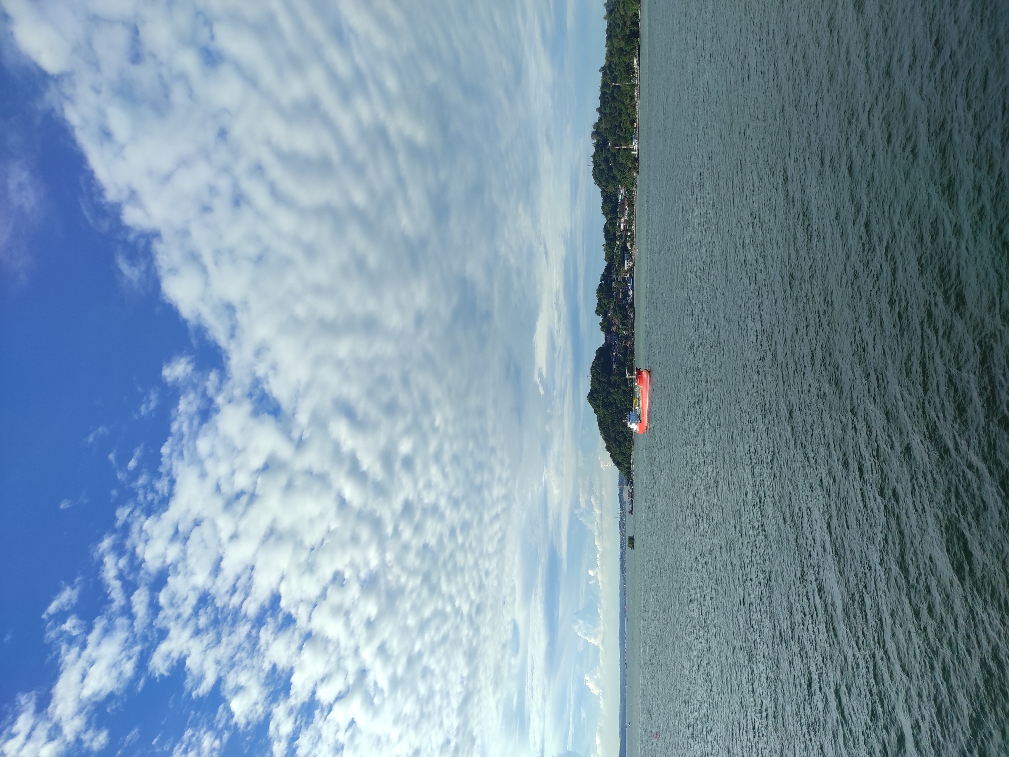 An eye-level, wide shot captures a tranquil seascape in Penajam, Indonesia, under a partly cloudy, bright blue sky. The foreground is dominated by the vast, dark blue expanse of the ocean, its surface rippled by gentle waves. In the mid-ground, a prominent bright red cargo ship sails across the water. Beyond the ship, a lush, green, tree-covered island stretches across the horizon, dotted with buildings and structures suggesting a coastal settlement. The sky above is a vibrant blue, adorned with scattered white, wispy clouds that form a textured pattern across the upper portion of the image. The overall scene conveys a sense of calm and vastness, typical of a coastal view.