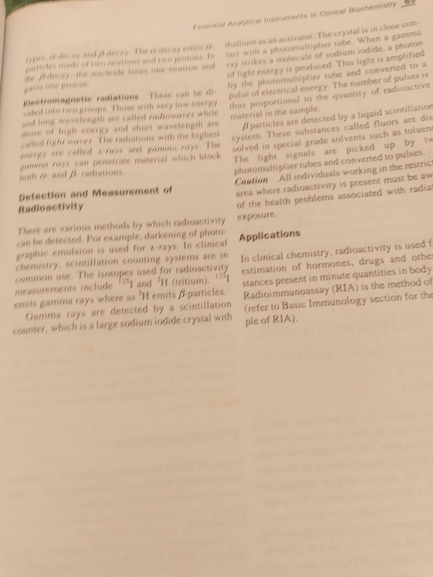 The image is a close-up of a page from a textbook, likely discussing analytical instruments in clinical biochemistry. The text is printed in black on a light-colored, slightly textured paper. The page is filled with dense paragraphs explaining various scientific concepts related to radioactivity, its detection, measurement, and applications.
The visible text covers topics such as alpha (α) and beta (β) decay, electromagnetic radiation (radio waves, light waves, X-rays, gamma rays), scintillation counting systems, and Radioimmunoassay (RIA). It details the processes involved in detecting radioactive particles and the use of specific isotopes like Iodine-125 and Tritium in measurements. The text also includes a caution about working with radioactive materials.
There are no people, objects other than the book itself, or a discernible scene or setting visible in the image. The lighting appears to be neutral, suggesting an indoor environment, likely a library or study area. The overall mood conveyed is academic and informative. There are no specific time-of-day or weather cues. The location context of Jalingo, Nigeria, is not directly inferable from the image content itself.