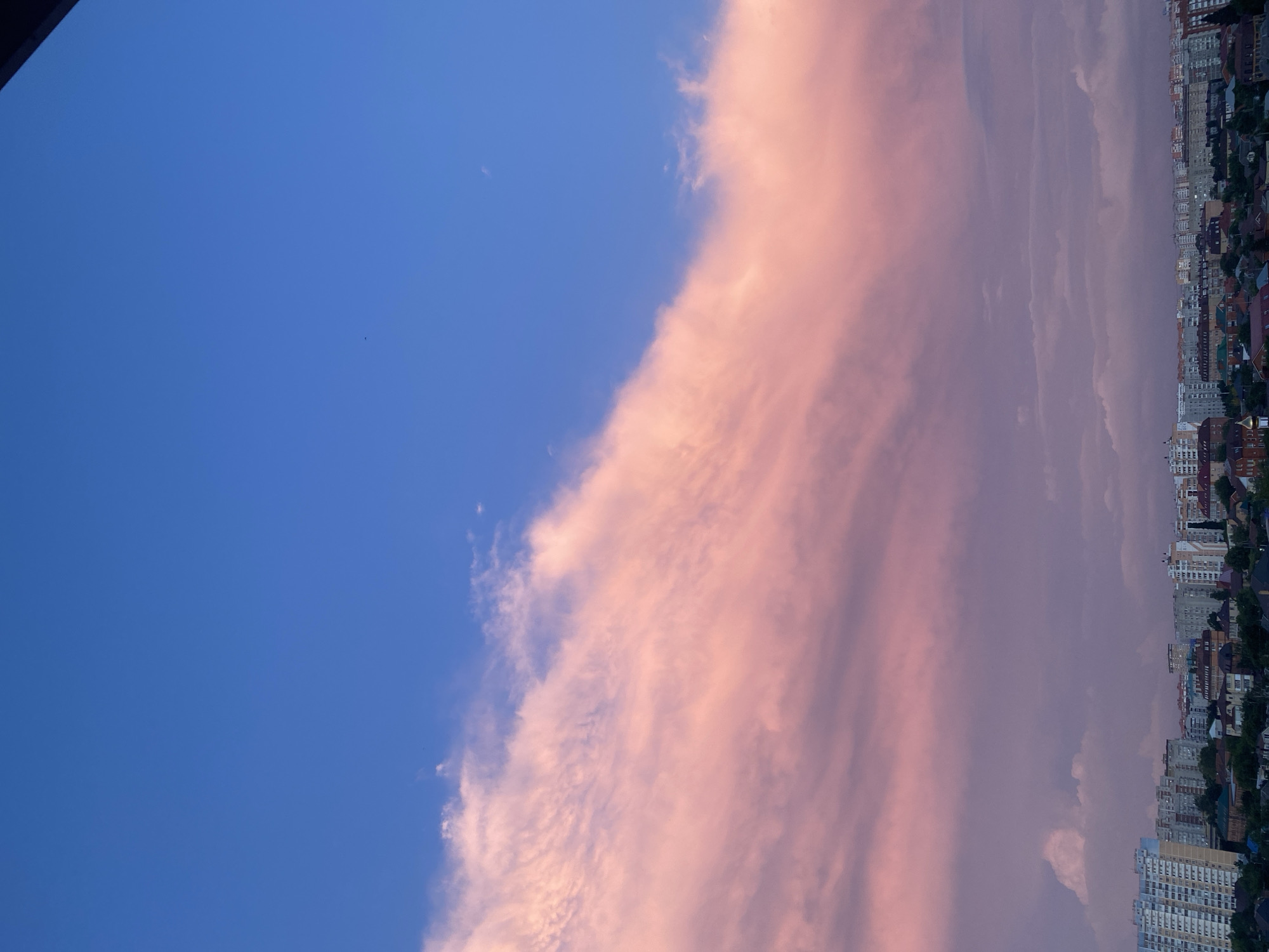 The image captures a cityscape at dusk, with a vibrant pink and blue sky dominating the upper portion. A large, illuminated cloud formation stretches diagonally across the frame, appearing as a soft, cotton-candy-like mass against the deep blue expanse. Below the sky, a dense collection of buildings forms the city's skyline. These structures vary in height and architectural style, with visible apartment buildings, smaller houses with pitched roofs, and some with darker, more industrial designs. Trees and greenery are interspersed among the buildings, suggesting a residential or mixed-use urban area. The lighting indicates either sunrise or sunset, with the warm hues of the clouds casting a gentle glow over the scene.