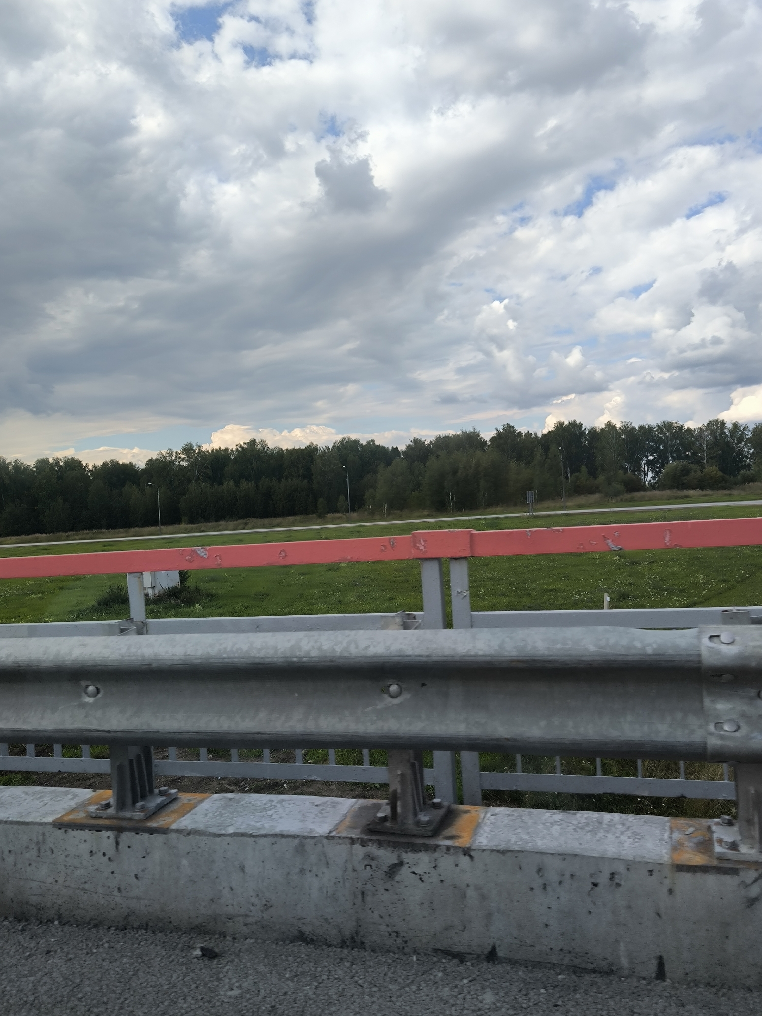 The image shows a guardrail in the foreground, with a concrete barrier beneath it. Beyond the guardrail, there is a green grassy field with some small white flowers. A red horizontal bar runs across the middle ground, supported by grey vertical posts. In the background, a dense line of trees forms a treeline. The sky is overcast with large, grey clouds interspersed with patches of blue. The scene appears to be located near Barlak, Russia, as suggested by the location tag. There are no people or animals visible in the image. The overall impression is that of a roadside view on a cloudy day.