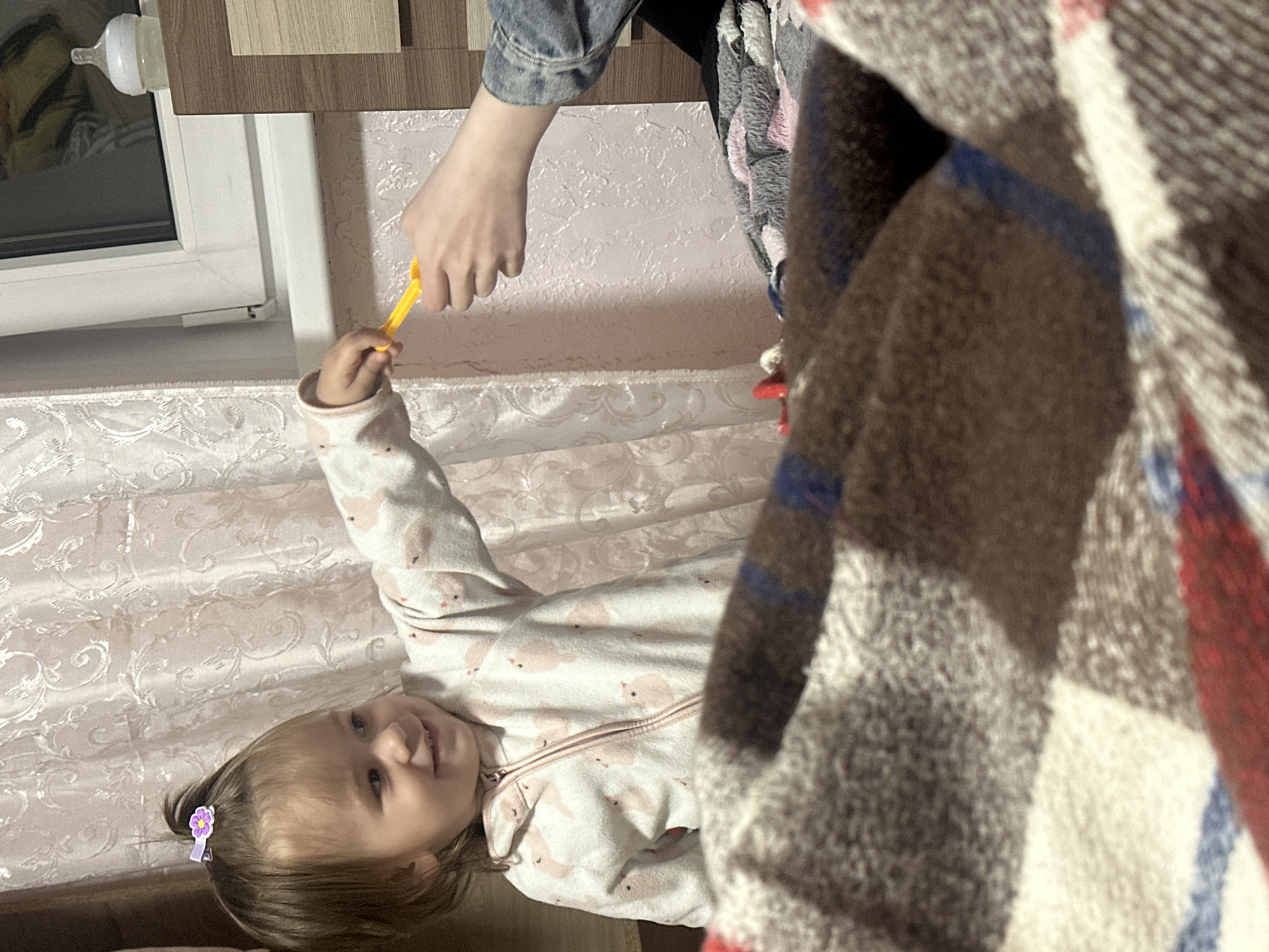 A young child lies on a patterned surface, possibly a bed, with a hand reaching towards her, offering a yellow object. The child is wearing a patterned jumpsuit, and her hair is adorned with a purple flower clip. A blanket with a plaid pattern is partially obscuring the view, and a window with a baby bottle attached to the frame is visible in the background. The location is Uva, Russia.