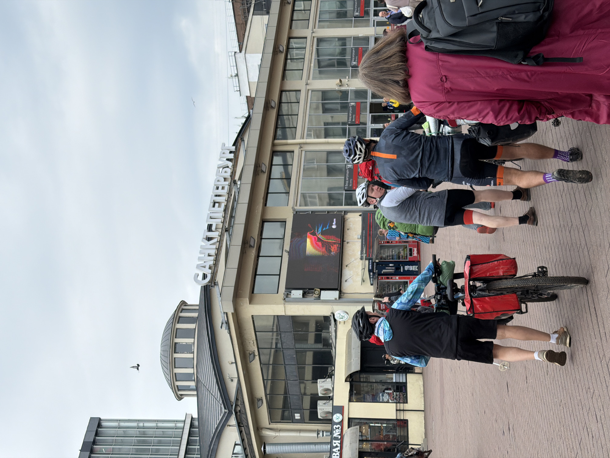 In front of the Saint Petersburg Glavny railway station, a group of people, some with bicycles, are gathered on a paved plaza. A man in athletic wear and a helmet, with a bicycle outfitted with red panniers, is seen from behind, walking towards the right. To his left, another cyclist in a grey jersey and helmet is visible. A woman in a bright pink coat and backpack stands in the foreground, facing away from the camera. The station's facade features large windows and signage, including the prominent inscription "САНКТ-ПЕТЕРБУРГ". The sky is overcast, and a bird is visible flying in the upper left quadrant of the image. The scene suggests a moment of pause or preparation for a journey, likely involving cycling.