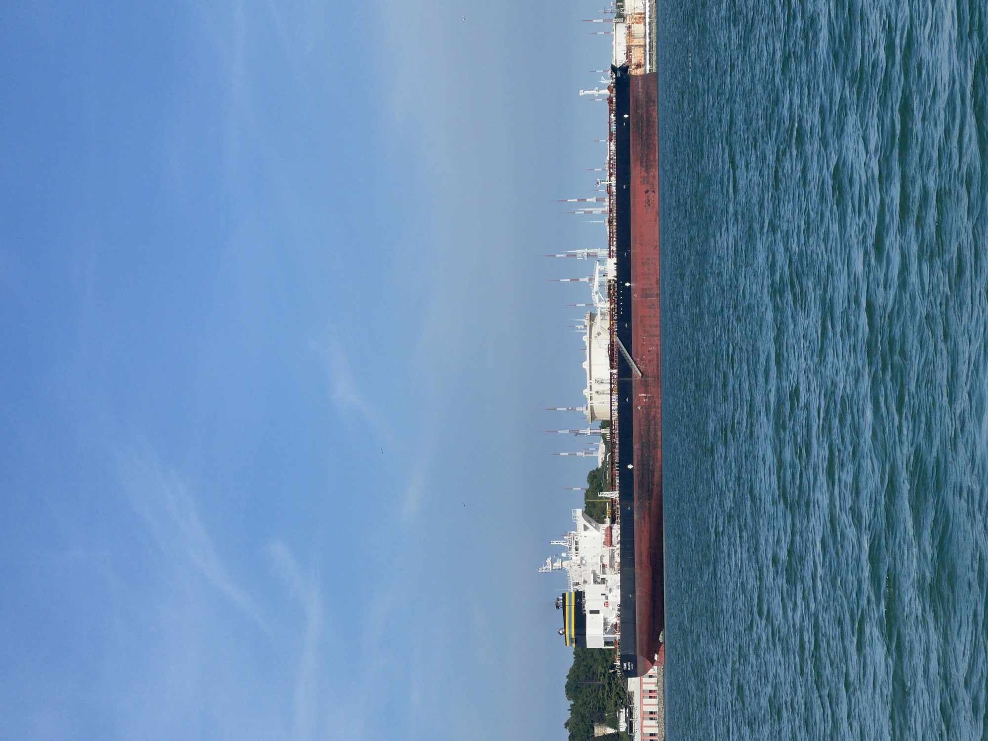 The image shows a large tanker ship docked at a port in Burgas, Bulgaria. The ship has a red-brown hull with white superstructure and is surrounded by choppy water. The sky is blue with some wispy clouds. In the background, there are industrial buildings and greenery visible near the coastline.