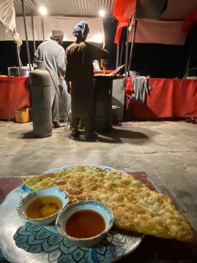 In Kandahar, Afghanistan, a dimly lit street scene unfolds at night. The foreground features a plate of fried dough, accompanied by two small bowls containing dipping sauces. Behind this food presentation stand two men, seemingly working at a food stall. The taller man on the left is wearing a light-colored outfit and hat, while the other man, positioned on the right, is clad in darker attire and a headwrap. Both appear to be preparing food. A propane tank is visible to the left of the food stall. The stall itself has a red covering, with several poles standing in the background.