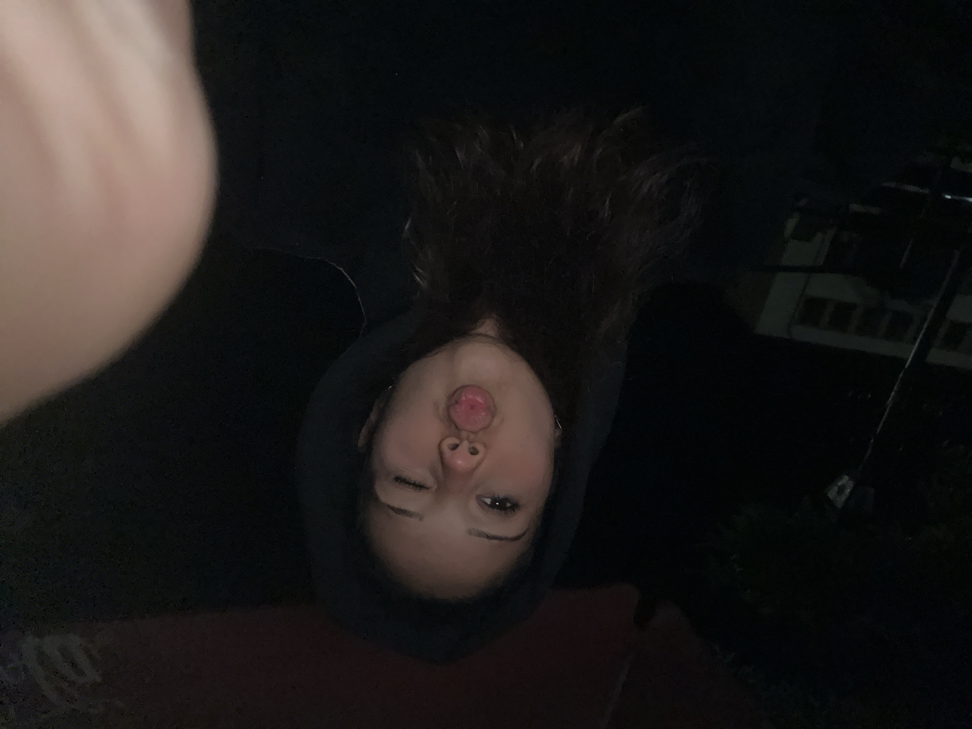 A young woman is pictured in a close-up shot from the chest up, wearing a black hooded sweatshirt. Her hood is up, obscuring the top of her head. She has long, dark, wavy hair that falls past her shoulders. She is winking with her right eye, and her lips are puckered, with her tongue slightly protruding. The background is dark, suggesting it is nighttime or an indoor setting with low lighting. To the left of the frame, a blurred image of a building with windows is visible. It is not possible to determine the specific location within the city of Zlatograd, Bulgaria, from the visual information provided.