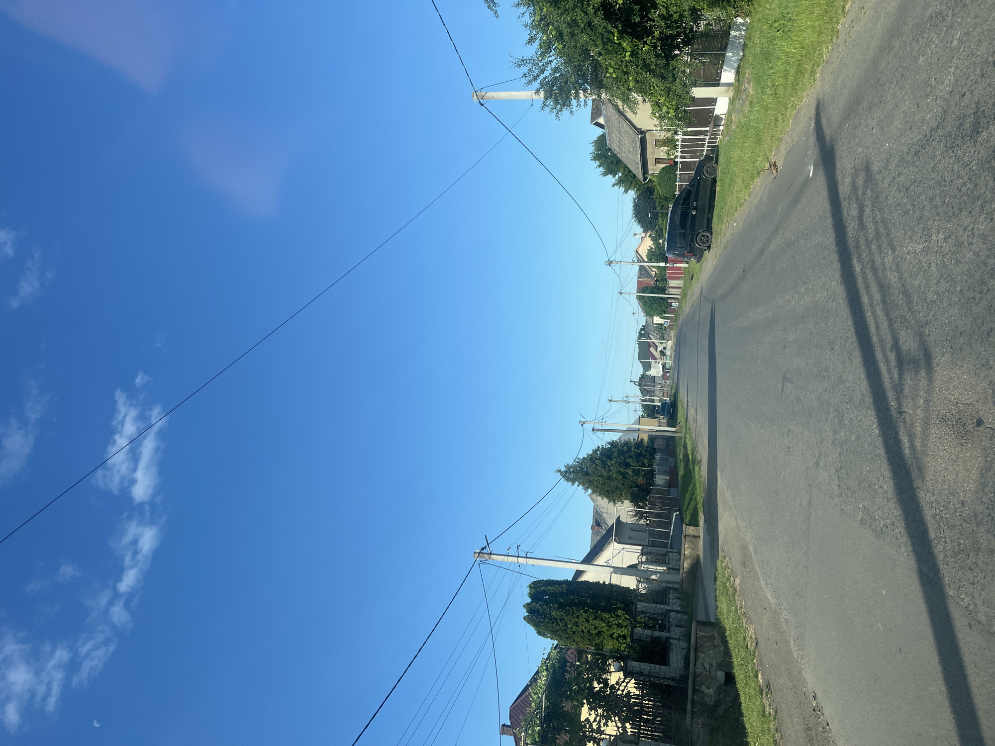 The image depicts a street in the village of Tarnivtsi, Ukraine, under a clear blue sky with a crescent moon visible. The perspective is from a vehicle driving down a paved road lined with houses and fences on both sides. Several utility poles with electrical wires are present along the street. On the left side, there are houses with red-tiled roofs and green trees. On the right side, a black van is parked near a fence. The road appears to be in a rural or suburban setting.