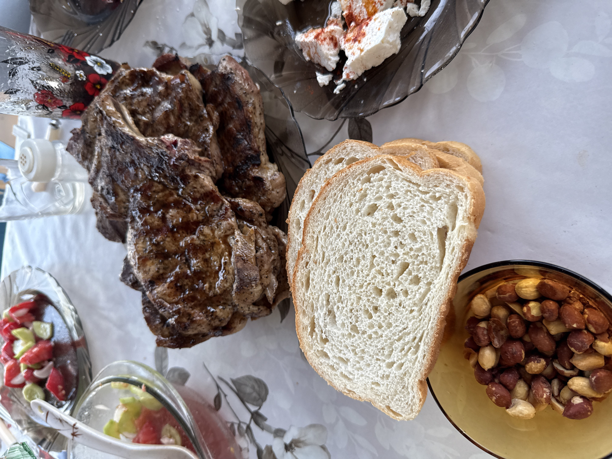 A close-up, eye-level shot captures a rustic meal spread on a white tablecloth with a floral pattern, possibly in Ravadinovo, Bulgaria. The main subject is a stack of grilled meat, likely steaks, with visible char marks and a glistening, browned exterior. To the left of the meat are slices of white bread, their porous texture evident. A small, amber-colored bowl filled with roasted peanuts sits in the foreground, partially obscured by the bread. To the right of the meat, a dark, wavy-edged plate holds a white cheese, likely feta or a similar soft cheese, sprinkled with red spice, possibly paprika. In the background, two glass dishes containing chopped tomatoes and cucumbers, dressed in a dark liquid, suggest a salad or appetizer. A glass with a decorative floral print, possibly holding a beverage, and a salt shaker are also visible in the background, hinting at a meal setting. The lighting is natural and diffused, creating soft shadows and highlighting the textures of the food. The overall impression is one of a hearty, casual outdoor meal.
