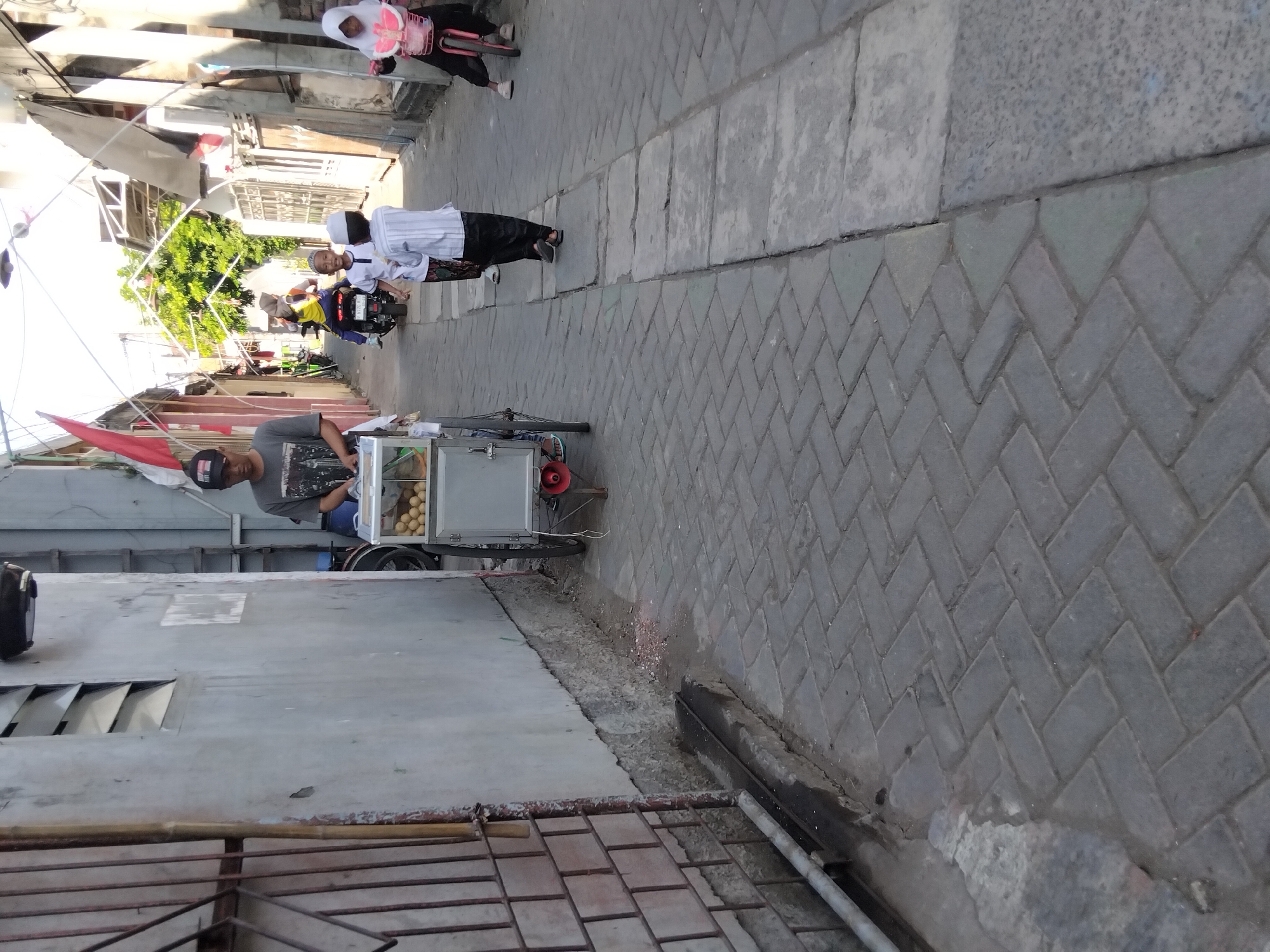 In Kepuh Kiriman, Indonesia, a narrow, paved alleyway is depicted under daylight. A man wearing a gray t-shirt and a black baseball cap pushes a mobile food cart. The cart, made of stainless steel, displays what appear to be small, round food items, possibly eggs, in a clear container. Two young boys in white shirts and black pants, one wearing a white cap, walk away from the camera down the alley. A young girl on a pink bicycle with training wheels rides in the same direction. Further down the alley, a person rides a motorcycle, and another individual, wearing a yellow shirt, is seated on the back. The buildings lining the alley are simple, with some featuring exposed wiring and a red and white Indonesian flag prominently displayed. The ground is paved with interlocking gray bricks, and a gray concrete structure runs along the left side of the alley, separating the paved area from the wall of a building. A metal grate covers part of this concrete structure. The overall scene suggests a typical day in a residential or commercial area of the town.