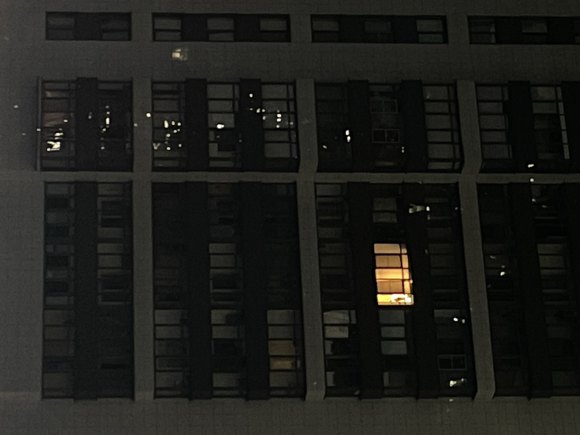 The image is a close-up, eye-level, exterior shot of a residential building at night in Sinyavino, Russia. The building appears to be made of concrete and glass, with multiple stories and numerous windows. Several windows are illuminated from within, suggesting that the apartments are occupied. The overall lighting is dim, consistent with nighttime conditions. The focus is on the architectural details of the building and the scattered lights from the apartments. There are no discernible people or specific objects visible outside of the building's structure and windows. The environment is urban, characterized by the monolithic apartment block.