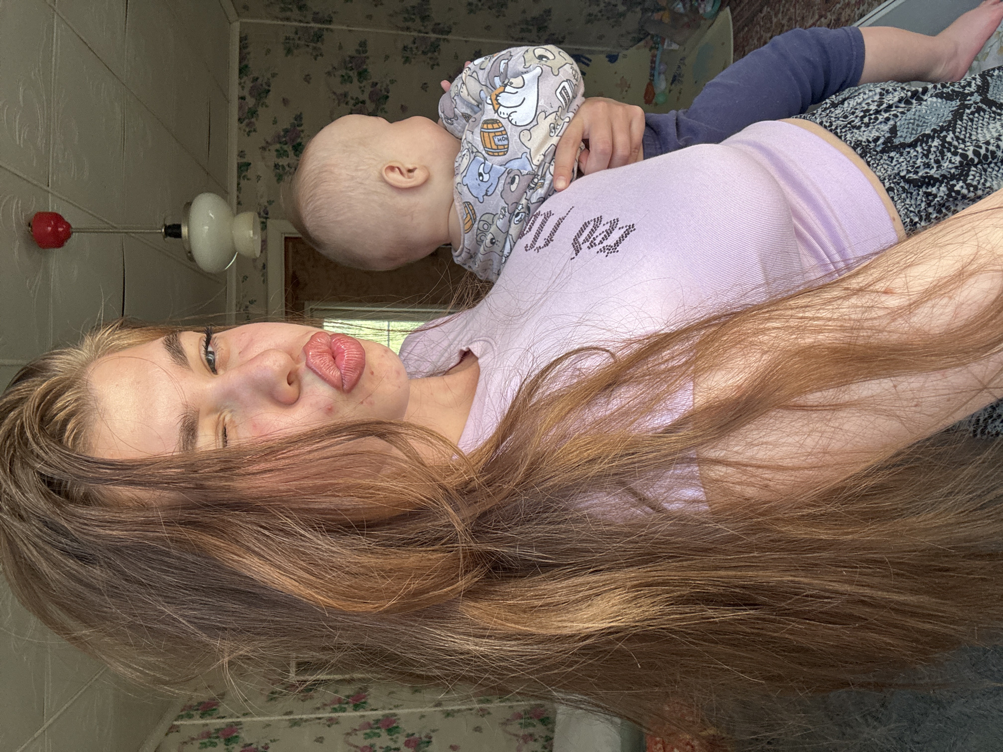 In a room with floral wallpaper and a textured ceiling, a young woman with long, auburn hair holds a baby. The woman is winking and puckering her lips for a selfie, her face partially obscured by her hair. She is wearing a light purple, ribbed crop top and patterned pants. The baby, facing away from the camera, is dressed in a gray onesie with a cartoon bear print and navy blue pants. A light fixture with a red decorative element hangs from the ceiling. The overall impression is a casual, indoor snapshot, likely taken in a residential setting in Meshchery, Russia.