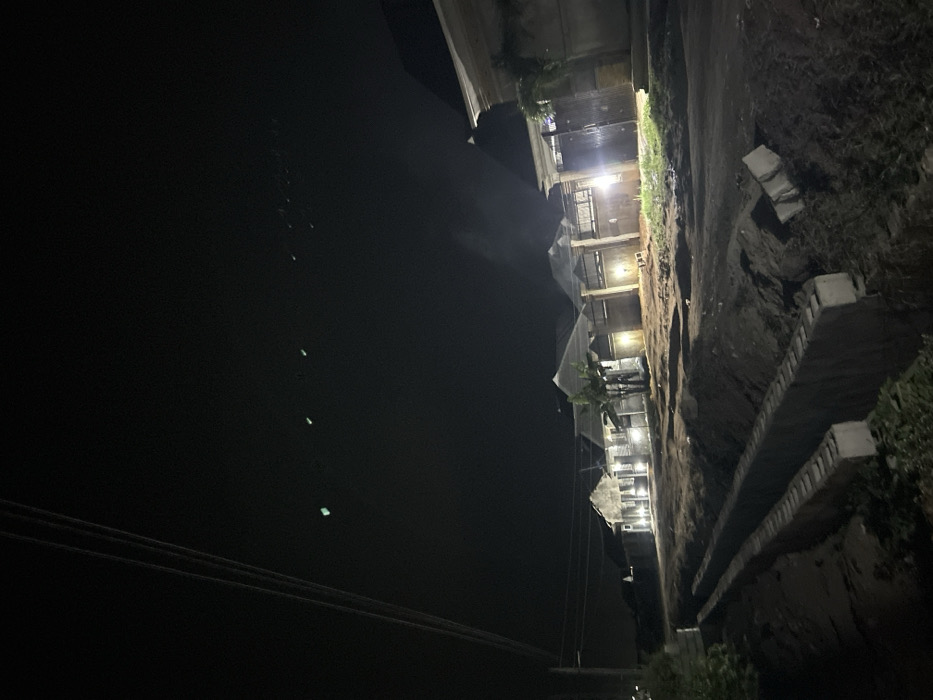 A nighttime scene shows a building illuminated by lights, adjacent to a steep, dirt slope reinforced with concrete. Power lines cut across the lower left of the frame, and a dark, undefined area occupies the majority of the left side. Small, indistinct green lights appear in the dark space. The setting has a gritty, somewhat desolate appearance.