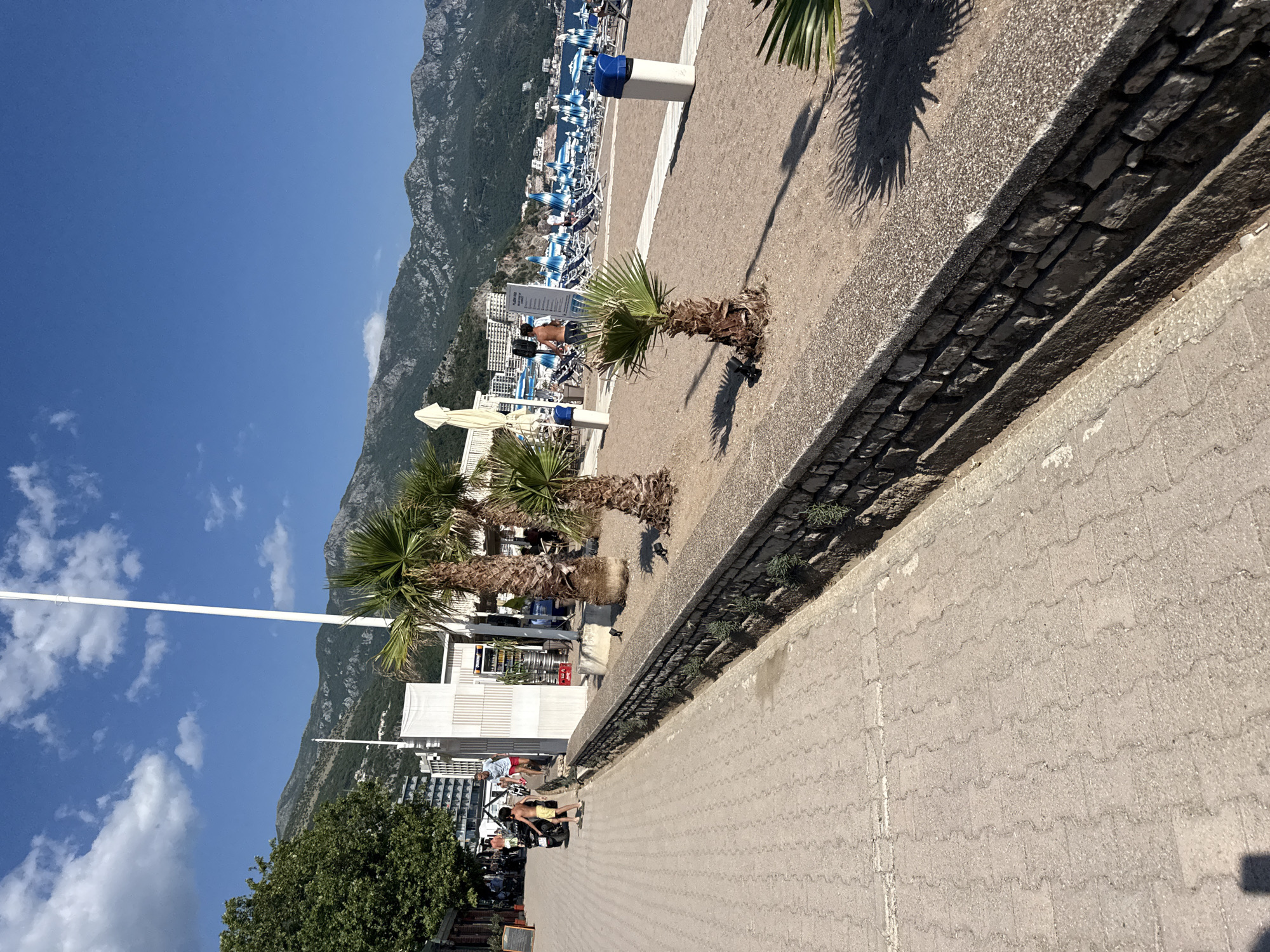 The image depicts a sunny day at a beachside promenade in Boreti, Montenegro. The foreground features a paved walkway with a low stone wall bordering a sandy area. Several palm trees are planted along the wall. In the background, the beach is lined with numerous blue and white striped lounge chairs and umbrellas. People are visible on the beach and promenade, some walking and others relaxing. Buildings, likely hotels, are situated behind the beach, and a mountain range forms the backdrop. The sky is clear blue with scattered white clouds.