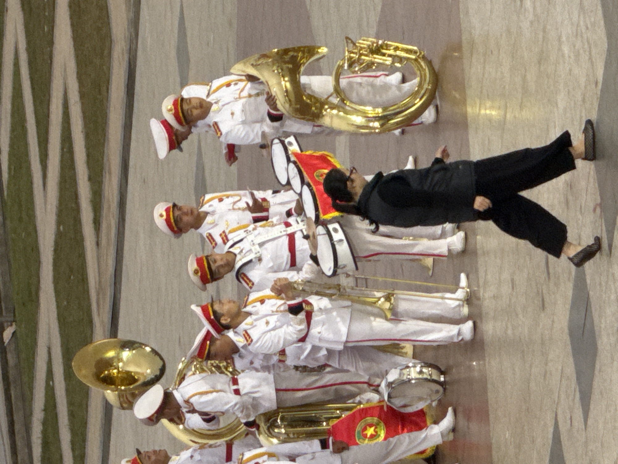 The image shows a marching band performing outdoors. The band members are dressed in white uniforms with red trim and red hats. They are playing various brass instruments, including tubas and trumpets, as well as drums. One person in the foreground is wearing black pants and a black jacket and is holding a red flag with a yellow star emblem. The ground appears to be paved. The scene suggests a formal event or parade. The lighting indicates it is daytime, likely under clear skies. Based on the provided context, the location is Da Lat, Vietnam. There is no visible text in the image.