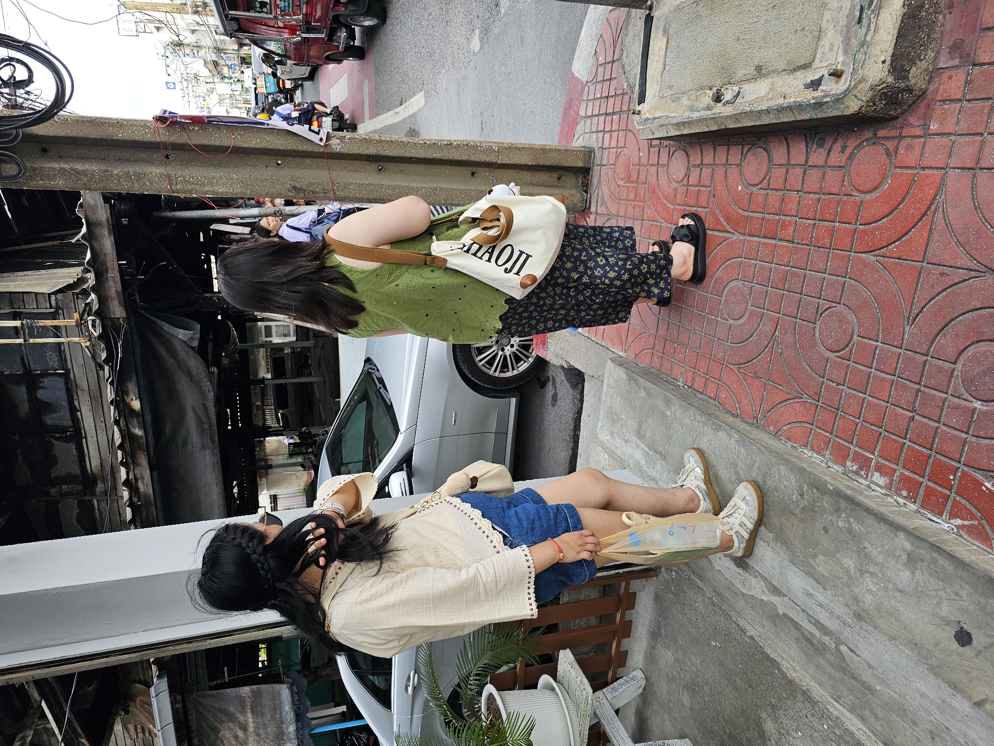 In Bangkok, Thailand, two women stand on a sidewalk next to a street. The woman on the left, wearing a cream-colored shirt and denim shorts, holds a plastic bag. The woman on the right, in a green top and patterned skirt, carries a canvas tote bag. A silver car is parked to their left, and a busy street with traffic, including a tuk-tuk, is visible to their right. The ground is paved with red tiles and concrete. Overhead, wires and a wooden structure suggest an urban environment.