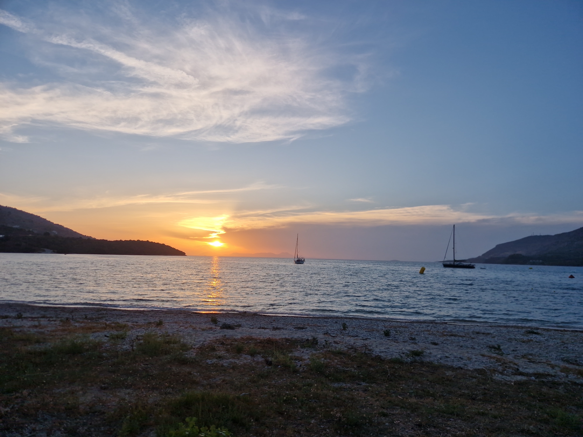 This is a landscape photograph of a tranquil beach scene at sunset in Plataria, Greece. The sun, partially obscured by a thin layer of clouds, casts a warm, golden-orange glow across the sky and reflects shimmering streaks on the gently rippling water. The horizon is defined by a dark, undulating silhouette of a distant coastline or island to the left, and a similar, but more mountainous, landmass to the right.
In the water, two sailboats are visible. The one on the left is a smaller white sailboat, appearing to be closer to the shore. The sailboat on the right is larger, darker, and further out, with its sail down. A yellow buoy floats in the sea between the two boats.
The foreground of the image is a beach composed of pebbles and sparse, dry vegetation. The beach slopes gently towards the calm water. The sky transitions from the warm hues of sunset near the horizon to a clear, soft blue higher up, with wispy white clouds scattered across the upper half of the frame. The overall atmosphere is peaceful and serene.