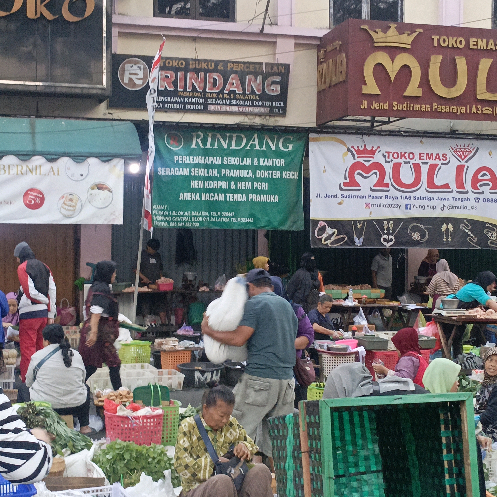 The image shows a street scene in Salatiga, Indonesia, likely a market or shopping area. There are several storefronts and stalls visible. One prominent sign reads "TOKO BUKU & PERCETAKAN RINDANG" (RINDANG Book & Printing Shop) with smaller text indicating its location in "PASAR AYAT 1 BLOK A No 8 SALATIGA." Another large green banner below this sign also displays "RINDANG" and lists "PERLENGKAPAN SEKOLAH & KANTOR SERAGAM SEKOLAH, PRAMUKA, DOKTER KECIL HEM KORPRI & HEM PGRI ANEKA MACAM TENDA PRAMUKA" (School & Office Supplies, School Uniforms, Scouts, Small Doctor's Office, KORPRI Uniform & PGRI Uniform, Various Types of Scout Tents). Further to the right, a sign for "TOKO EMAS MULIA" (MULIA Gold Shop) is visible, with the address "Jl. Jend Sudirman Pasaraya 1 A3 Salagita Jawa Tengah."
In the foreground, several people are present, some appearing to be vendors and others customers. There are many baskets and crates filled with goods, including what appear to be vegetables and other produce. One person is carrying a large white sack. Another person is sitting and appears to be sorting through items in a bag. The overall scene suggests a busy commercial environment with a mix of shops and street vendors. The architectural style of the buildings and the signage are typical of Indonesia.