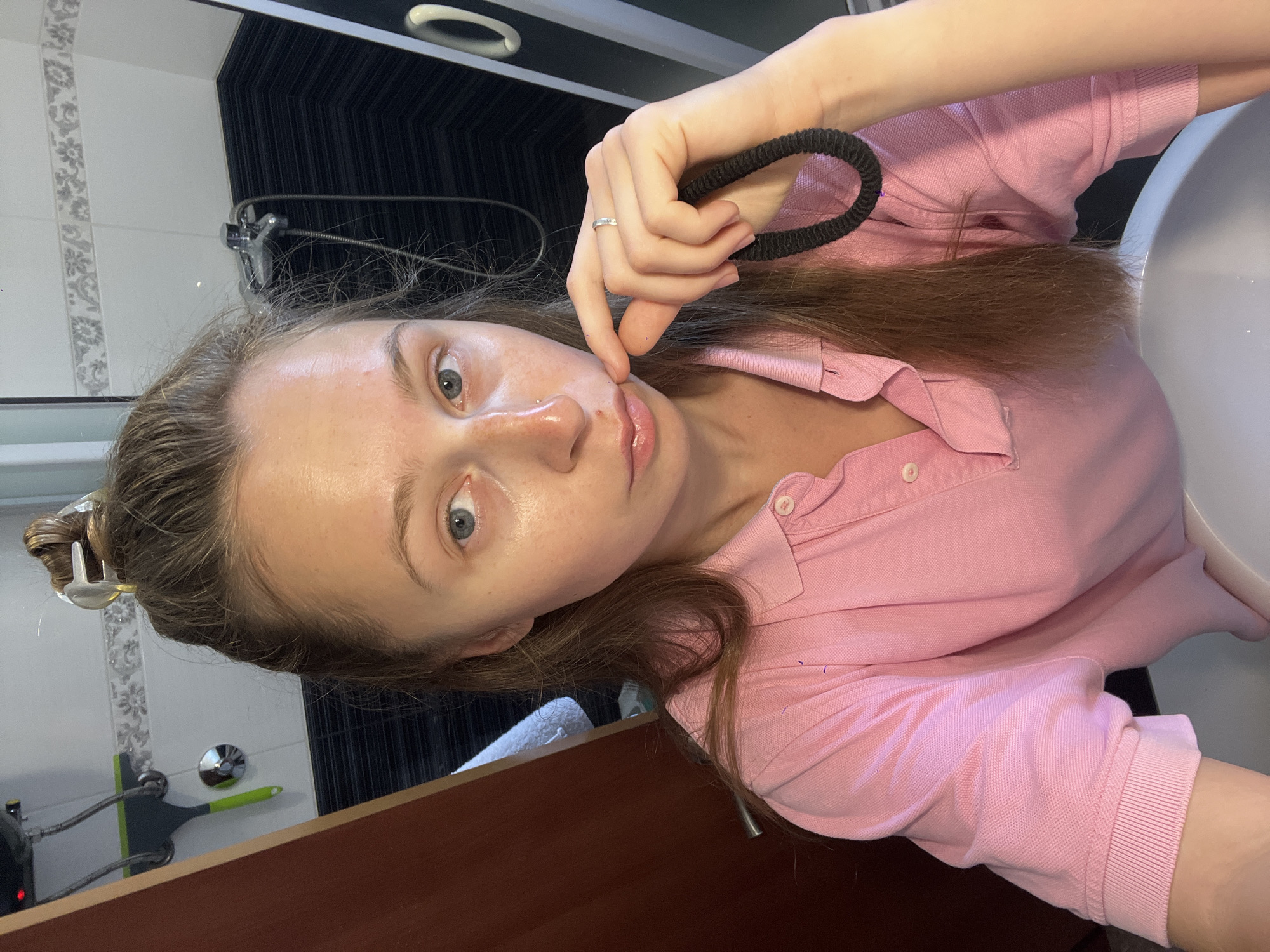 A young woman is pictured in a bathroom. She is fair-skinned, with brown hair partially pulled back with a clip. She is wearing a pink polo shirt and holding a black hair tie in her hand. Her left index finger touches her left cheek. She is wearing a ring on her left ring finger. The bathroom includes a sink, a mirror, and a dark-tiled wall with a shower head. There is also a brown cabinet below the sink. The location is likely a hotel or home bathroom and is not possible to discern as being specifically within the city of Bečići, Montenegro.