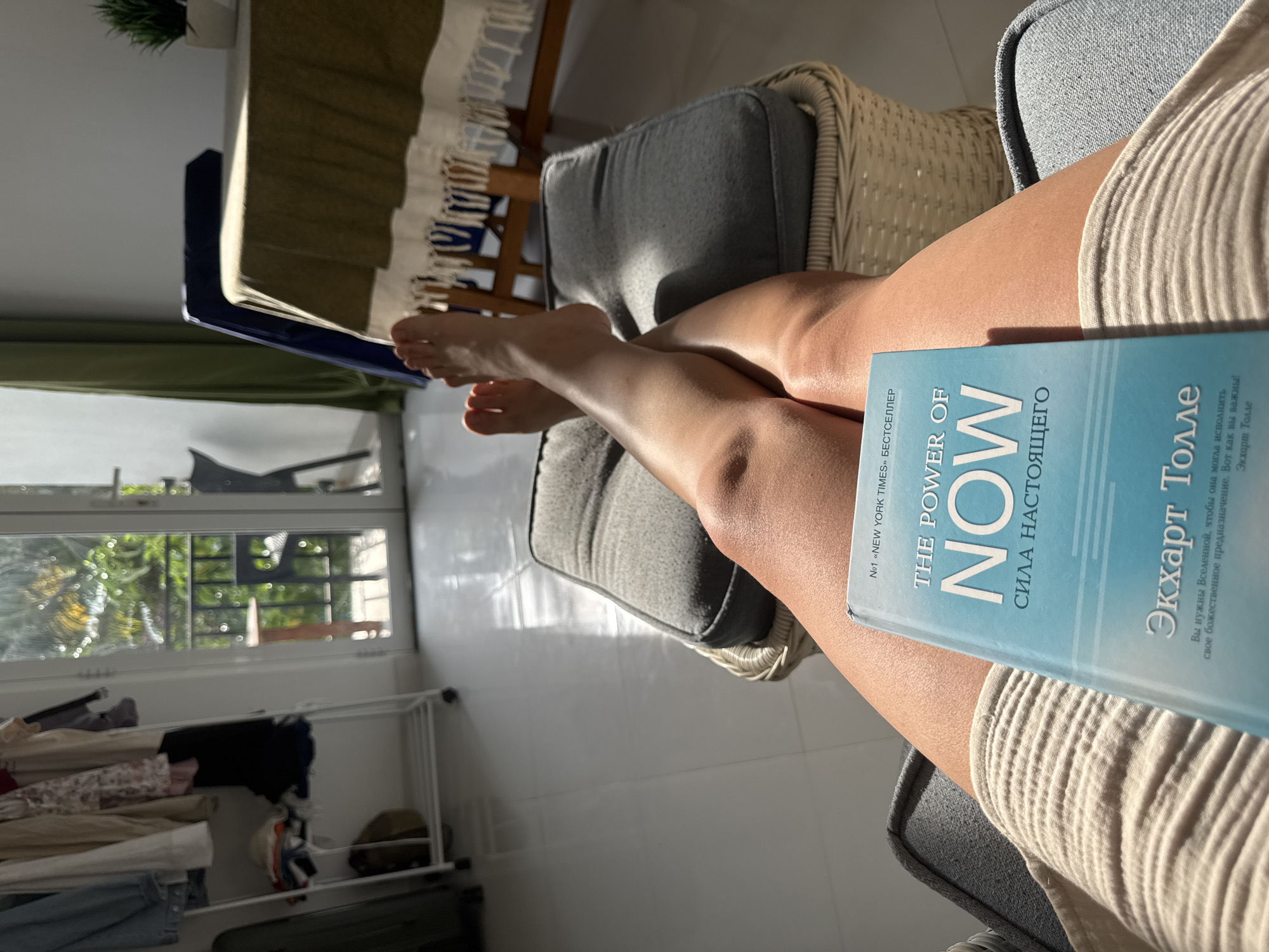 Here is a matter-of-fact description of the image:
The image presents a first-person view of a person relaxing indoors, likely in a residential setting. The individual is seated with legs extended, and a book titled "The Power of Now" by Eckhart Tolle rests on one of the legs. Visible beyond the person are elements of an interior space, including a window offering a glimpse of greenery outside, suggesting a garden or natural environment. A clothes drying rack is visible in the background. The lighting appears to be natural daylight. There are no indications of the specific location of Thalaramba, Sri Lanka, in the image.