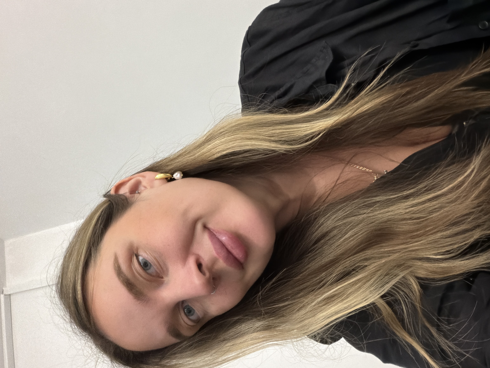 A close-up, tilted portrait captures a young woman looking directly at the camera with a slight smile. Her head and upper torso are visible, with her long, multi-toned blonde and light brown hair flowing across the frame, partially obscuring her black shirt or blouse. Her left shoulder and chest are visible, revealing a delicate, gold-toned chain necklace.
The woman has fair skin, blue eyes, and naturally defined eyebrows. She wears minimal makeup, with a focus on her lips which appear plump and a natural pink. Notable facial features include a small hoop nose ring on her left nostril. Her left ear is adorned with two gold-toned earrings: one is a small hoop or cuff with a white pearl, and another simpler design is also present.
The background is a plain, light-colored wall, suggesting an indoor setting. The image is significantly rotated clockwise, giving a dynamic, casual feel to the portrait. There is no visible text. The location context provided is Moscow, Russia.