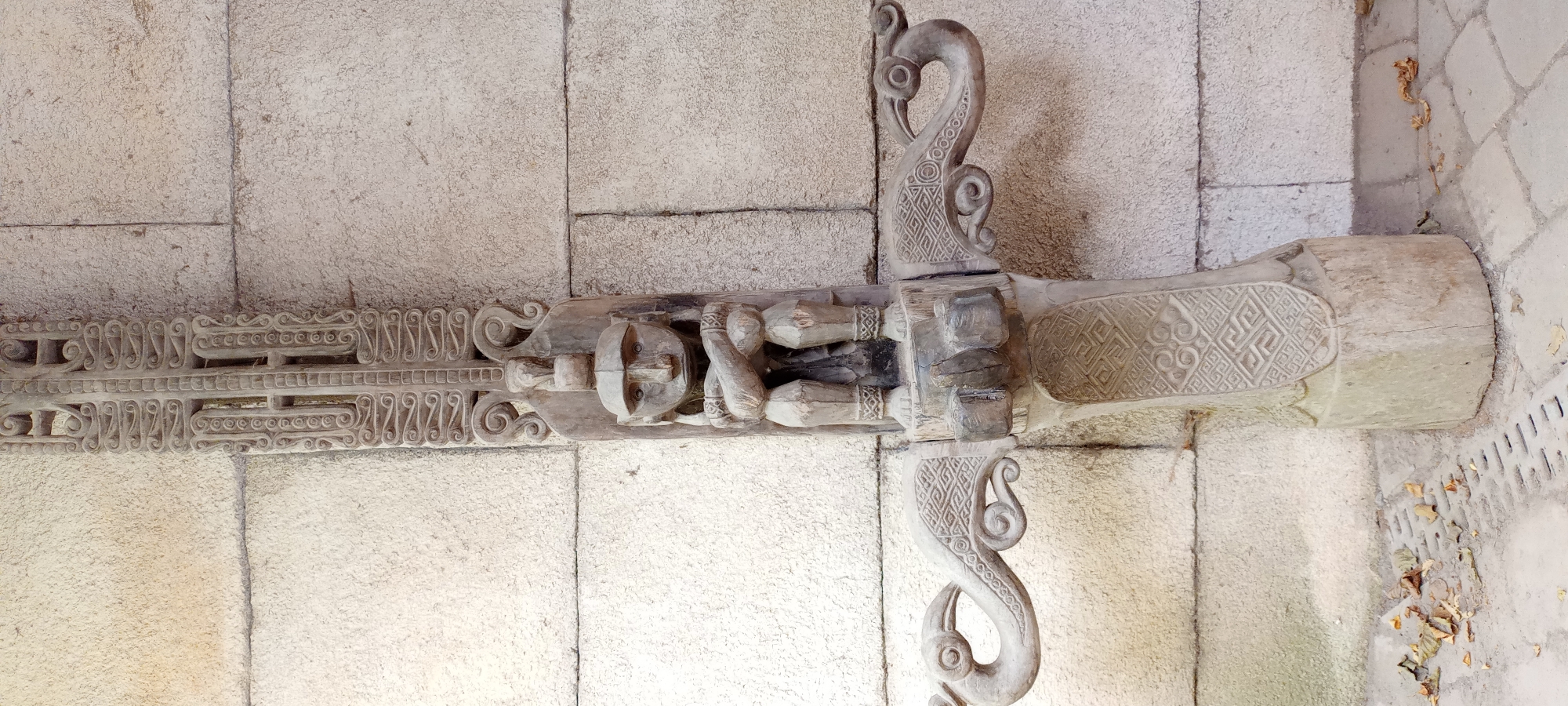 The image shows a close-up, eye-level, vertical shot of a weathered wooden carving mounted on a stone wall. The carving is ornate, featuring a seated figure with crossed arms and a distinct, angular face. Above the figure, the carving continues upwards with intricate geometric patterns and scrollwork. On either side of the figure, extending outwards horizontally, are carved elements resembling stylized birds or dragons. The lower portion of the carving also displays geometric patterns. The wooden object is attached to a textured stone wall, which appears to be made of large, light-colored blocks. In the foreground, there is a paved area with some fallen leaves and a metal grate. The overall impression is of an old, artistic installation. This specific carving is located at the "Haus der Begegnung" (House of Encounter) in Gelsenkirchen, Germany.