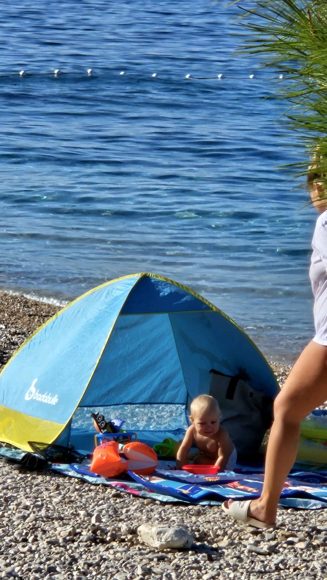The image depicts a sunny day on a pebbly beach in Općina Tučepi, Croatia. In the foreground, a blue and yellow beach tent is set up. Inside and near the tent, a baby is sitting on a patterned blanket, wearing a diaper and a red plastic bowl in front of it. Several beach toys, including an orange inflatable ring, are scattered on the blanket. To the right of the tent, a person's legs and feet are visible, wearing a white sandal. The background shows the calm blue water of the Adriatic Sea, with a line of buoys visible in the distance. A palm frond is partially visible in the upper right corner, suggesting lush vegetation nearby. The scene conveys a relaxed atmosphere of a family enjoying a day at the beach.