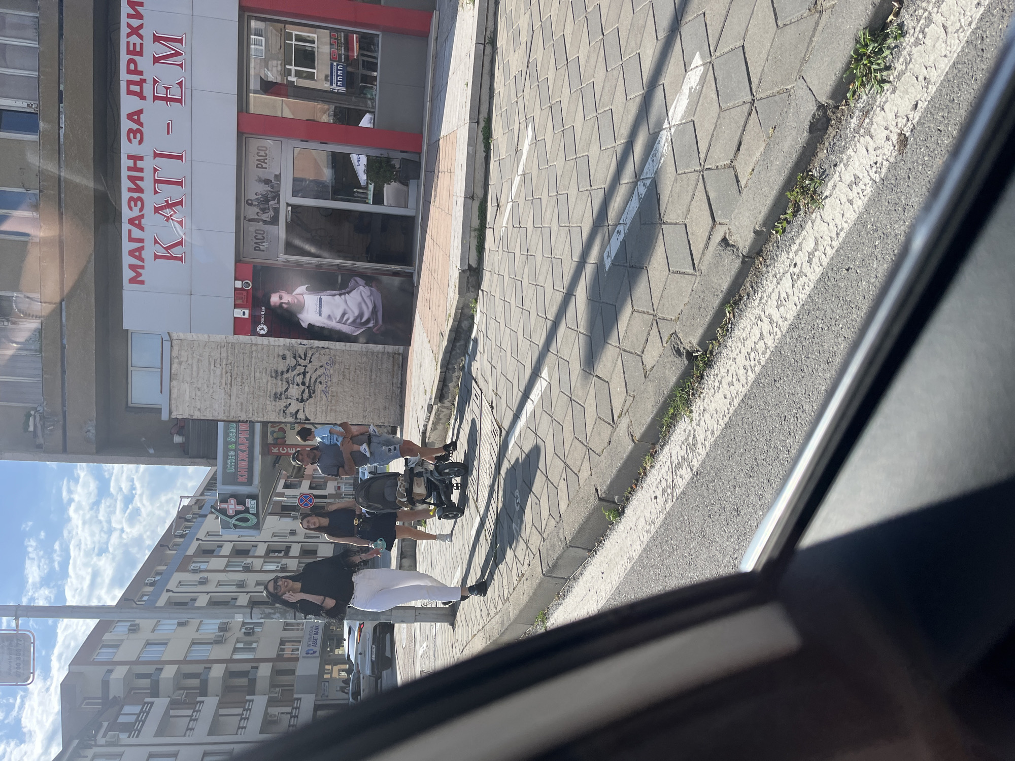 This image captures a daytime urban street scene in Blagoevgrad, Bulgaria, specifically near the "KATI-EM" clothing store and "International Asset Bank." The shot is taken from inside a vehicle, with the car window frame visible in the foreground, partially obscuring the lower left and bottom of the image. In the mid-ground, a group of four individuals is walking on a sidewalk paved with interlocking grey hexagonal tiles. From left to right: 1. A woman with long dark hair, wearing a black long-sleeved top, white pants, and black shoes. She is wearing sunglasses and is actively speaking on a mobile phone held to her right ear. 2. Another woman with long dark hair, wearing a dark dress or skirt and top, with a dark belt. She is pushing a dark-colored, three-wheeled stroller, which contains a light-colored plush item or blanket. She is holding a light blue bottle or cup in her left hand. 3. A man with a shaved head, wearing a grey t-shirt, dark shorts, and white sneakers. He is carrying a small child in his arms on his right hip. 4. The child being carried is wearing a light blue two-piece outfit with white stripes on the shorts. Behind the pedestrians, a white SUV is visible on the street, and other vehicles are parked further down. On the left, a multi-story residential building with numerous balconies and air conditioning units is prominent. Below it, the "INTERNATIONAL ASSET BANK" logo is visible. A light pole stands between the bank and the pedestrians. Further down the street, a large green sign with a red medical cross and the Bulgarian word "ШЕСТ" (Six) is attached to a building, below which is text indicating "Office & School КНИЖАРНИЦА" (Bookstore). On the right side of the image, a commercial building features a large sign that reads "МАГАЗИН ЗА ДРЕХИ" (Clothes Shop) in red Cyrillic letters, followed by "КАТИ – ЕМ" also in red. The storefront windows display "PACO" branding and a large poster of a man in a white sweatshirt. The street in front of the building has a faded white pedestrian crossing line and a continuous white line marking the edge of the curb. Distinct shadows are cast on the sidewalk, indicating bright sunlight from the left. Patches of weeds and grass are growing in the cracks along the curb and in the unpaved area between the curb and the main road. The